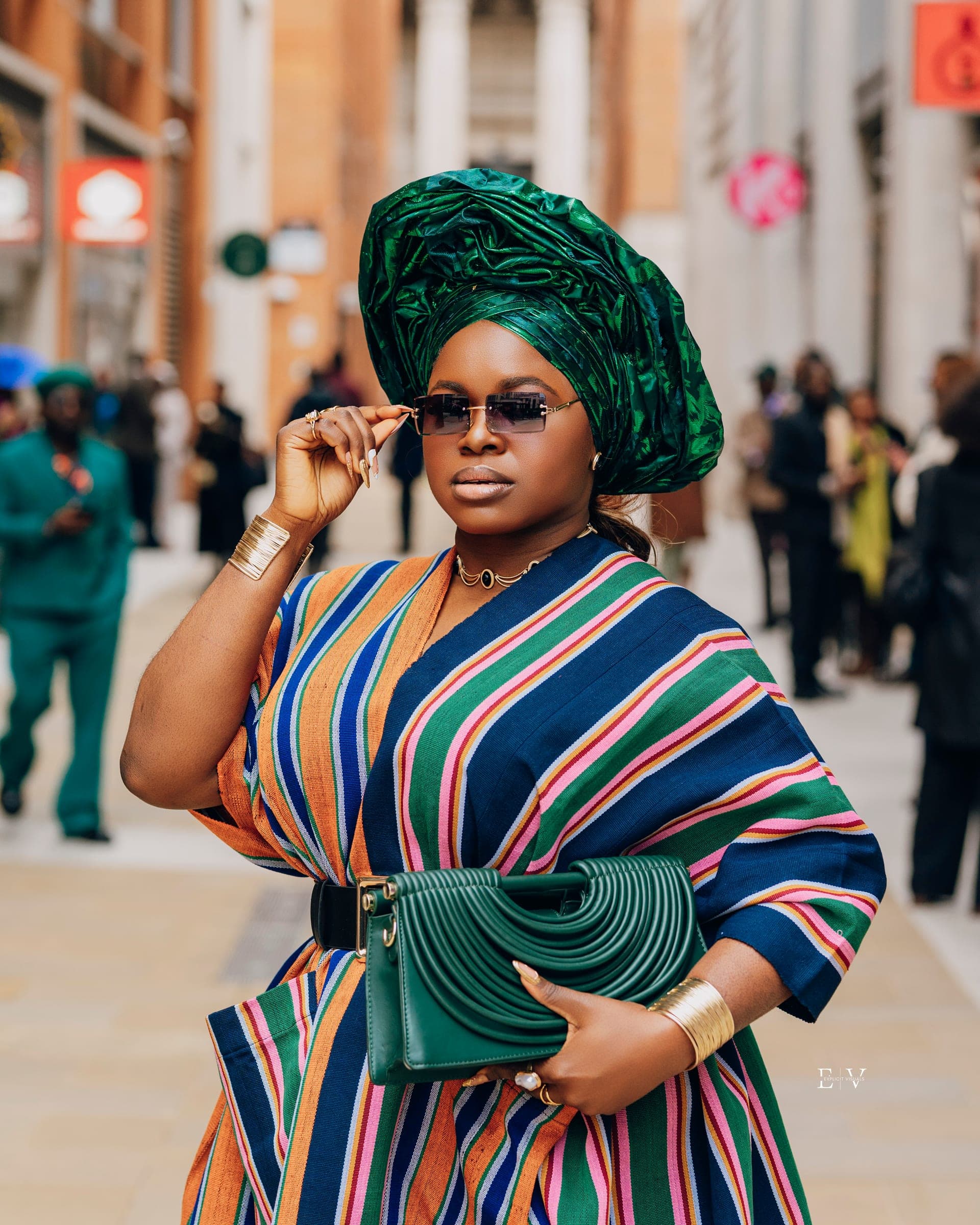 A closer frame of a green gele over sunglasses, a navy, pink and orange striped wrap, gold cuffs on both wrists and a green leather clutch held in front.