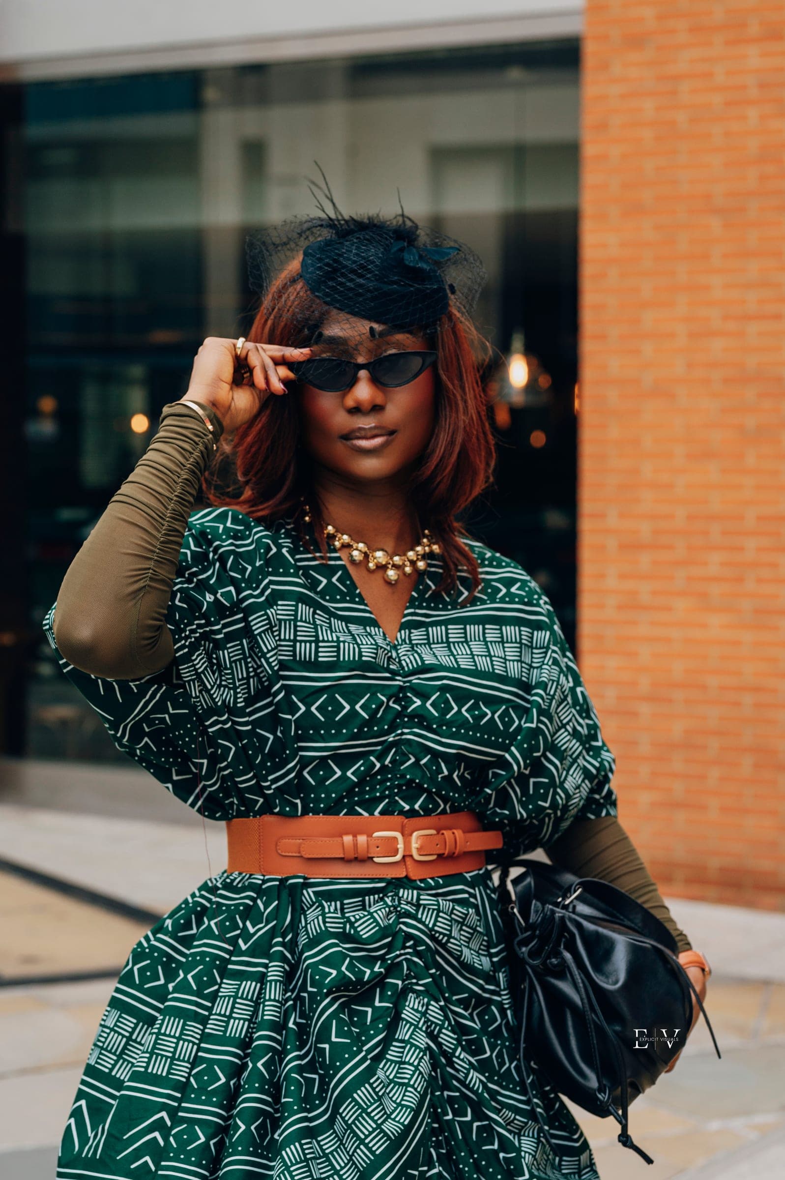 A woman in a black birdcage fascinator and cat-eye sunglasses lifting one hand to the frames, in a green geometric print dress with a tan waist belt.