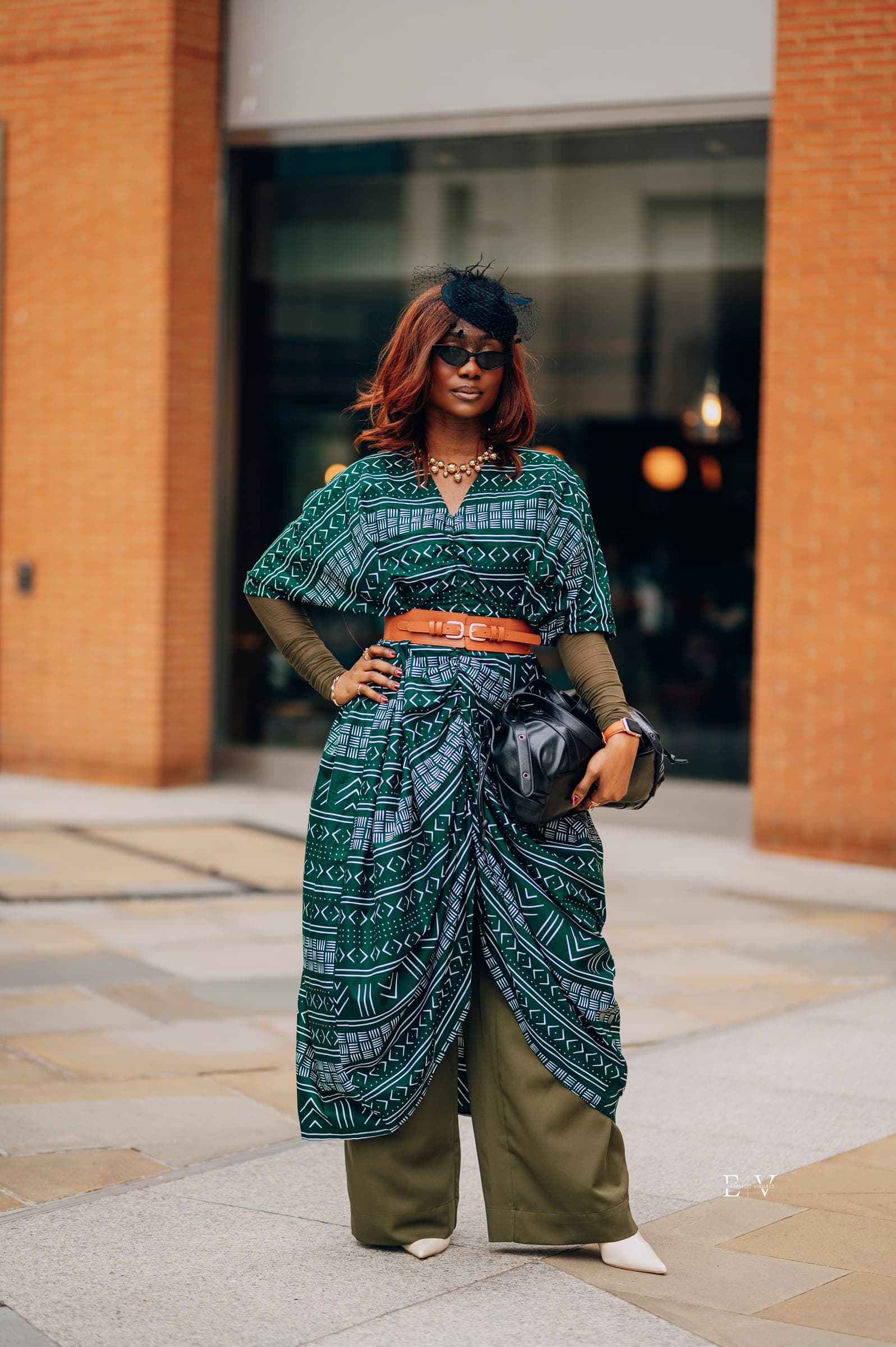 The same outfit full length — the printed wrap skirt knotted over wide olive trousers, a black leather bag in her hand, a brick wall behind.