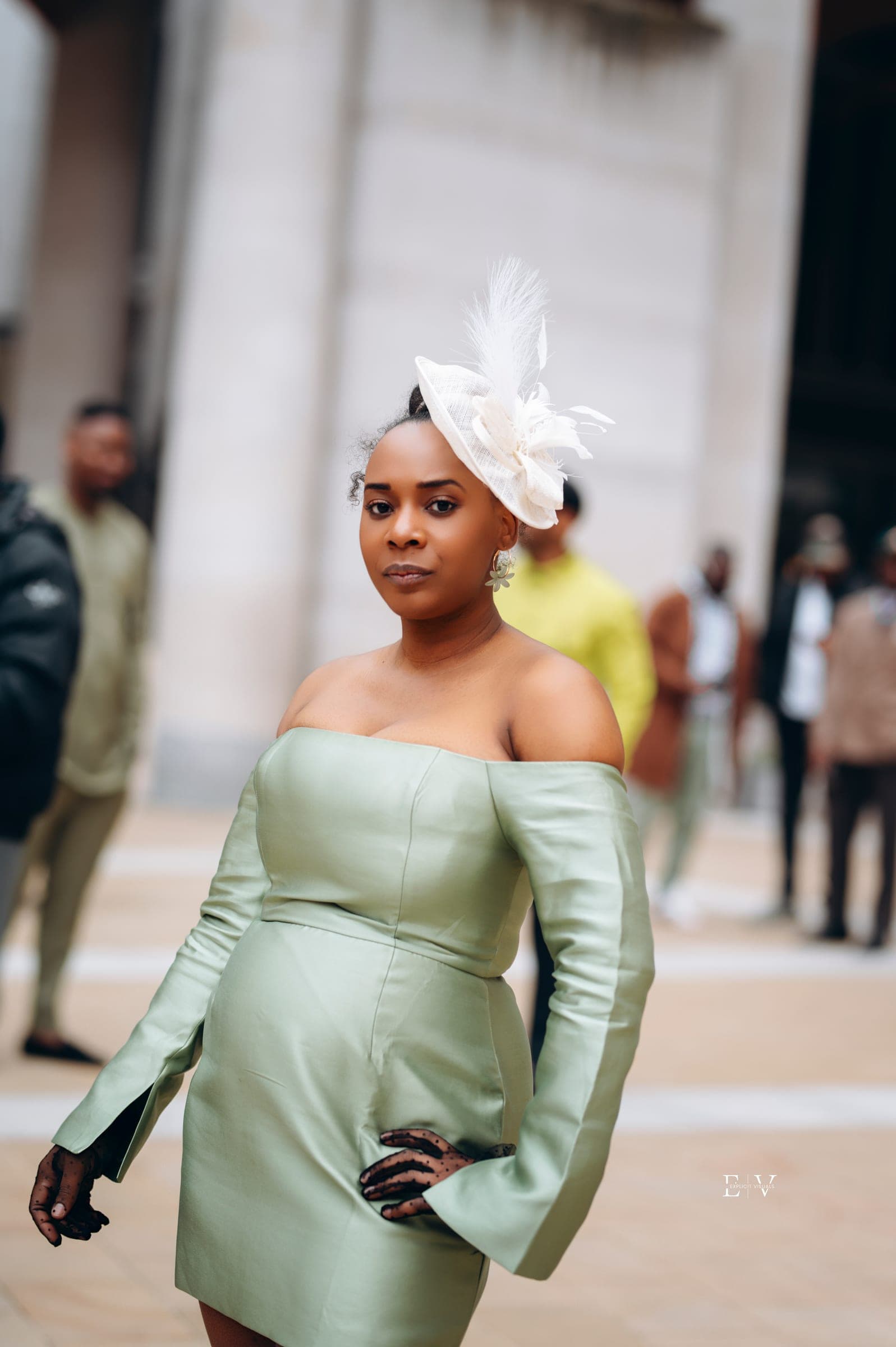 A woman in a sage green off-the-shoulder mini dress with long fitted sleeves and a cream feather fascinator, standing in the square.