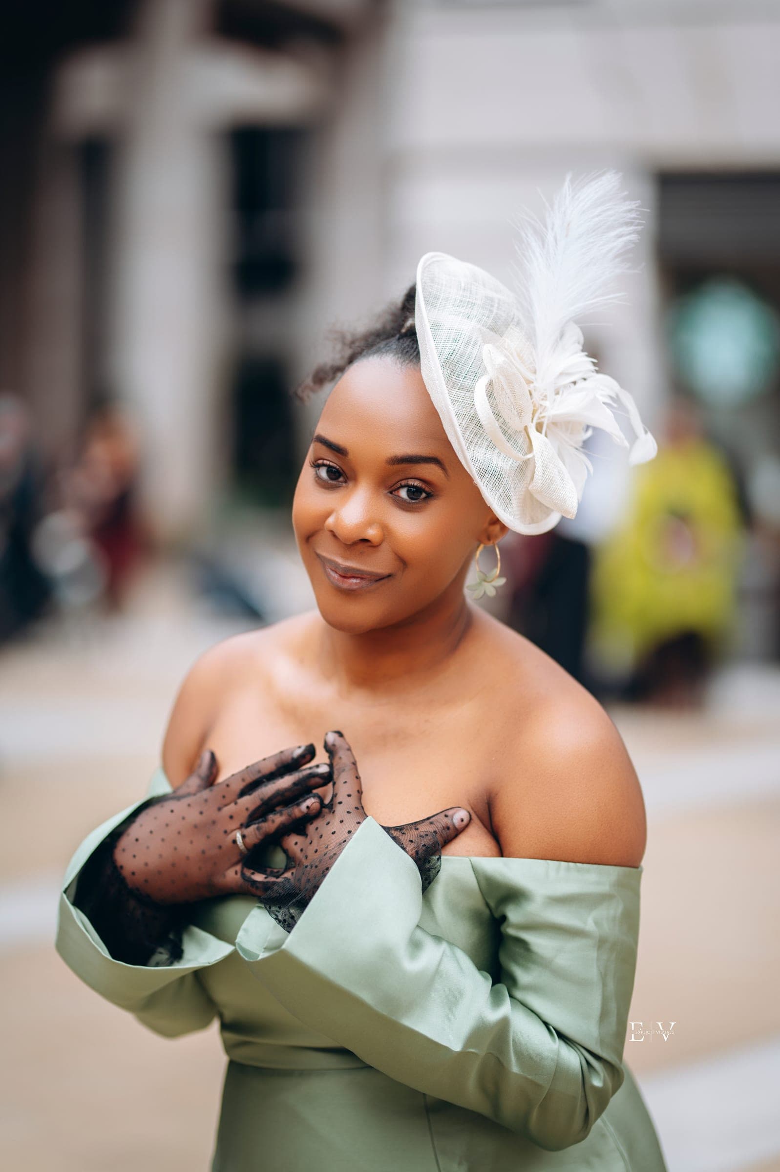 A close portrait — both hands crossed at her chest in sheer black polka-dot gloves, the feathered fascinator tipped over her hair.