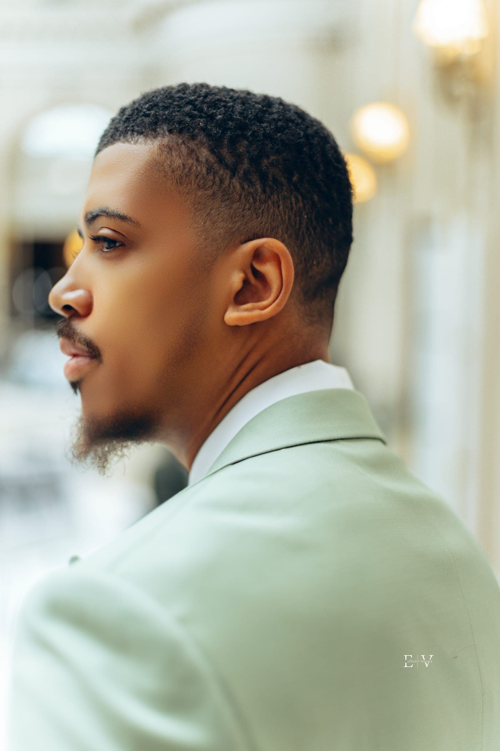 The same suit in profile, a tight frame on his face and shoulder, warm hanging lights out of focus down the hall.