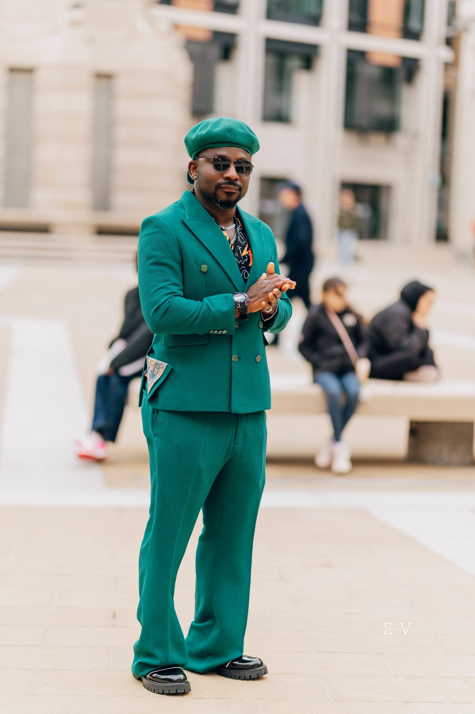 A man in an emerald green suit and matching flat cap over a printed shirt, gold chains at his neck, adjusting a ring as he looks to camera.