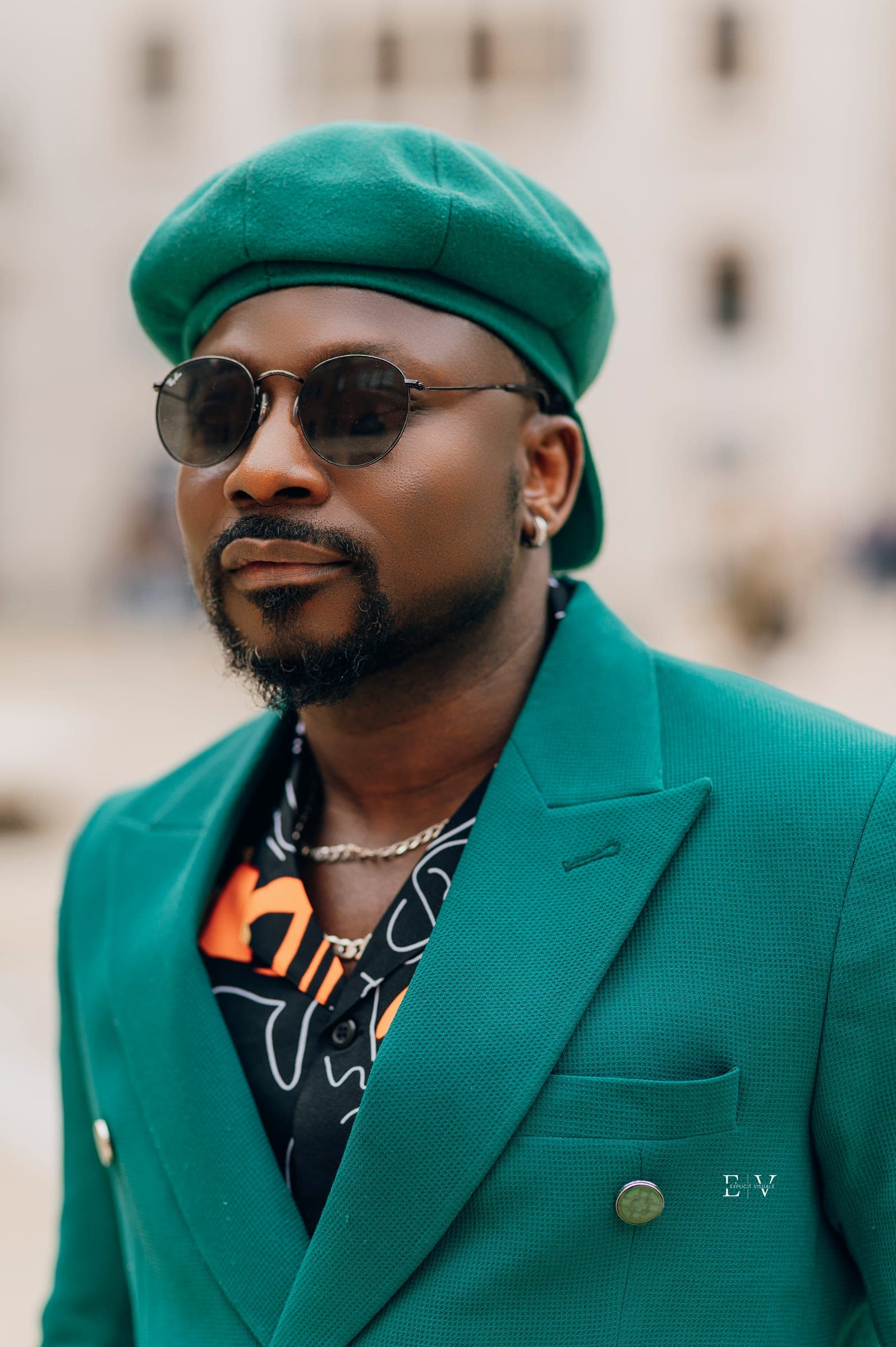 A close portrait of the same man — round dark sunglasses, green cap, the emerald lapel and gold buttons filling the lower frame.
