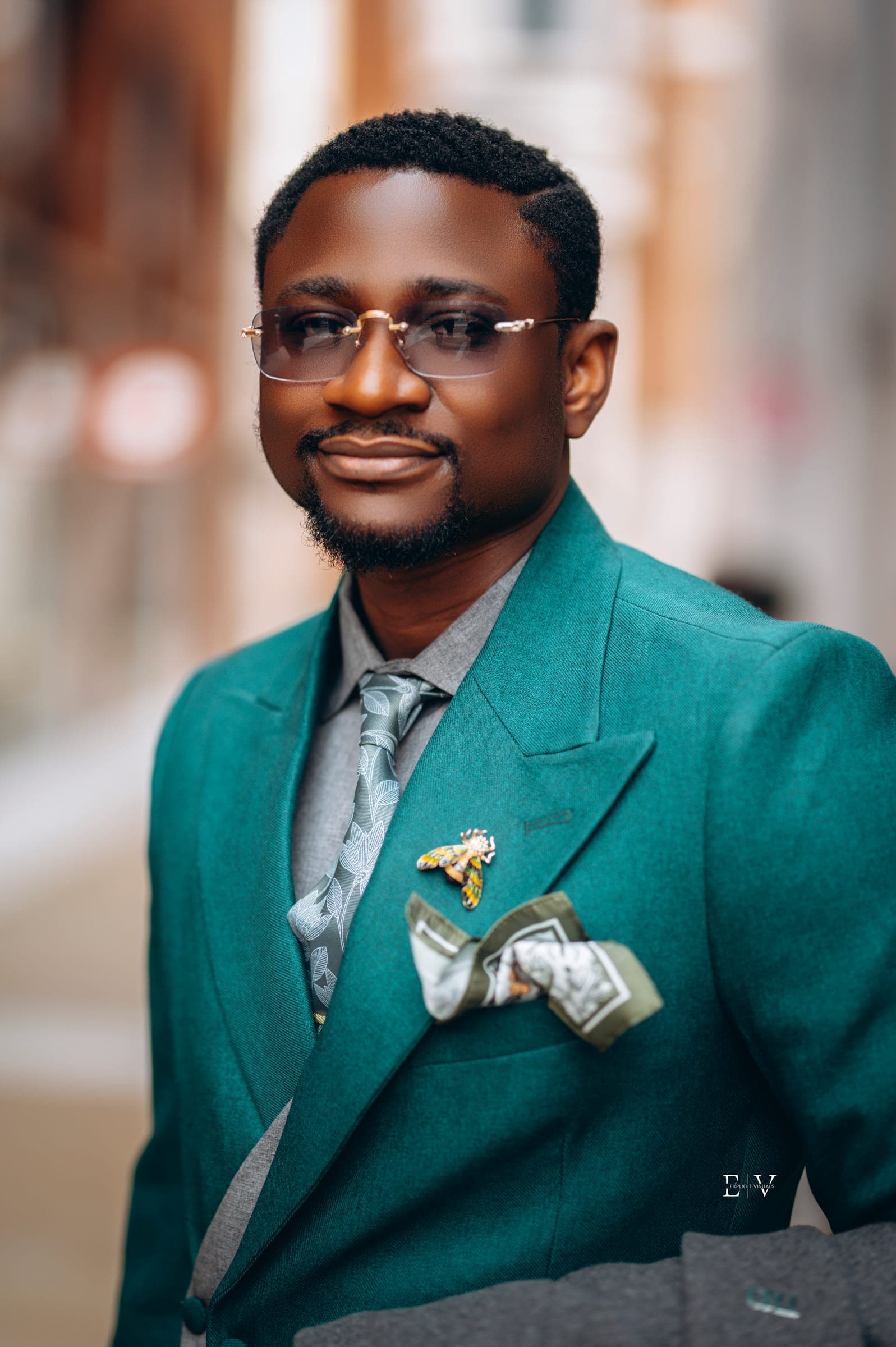 A close portrait of a man in glasses and a teal blazer over a grey waistcoat, a gold lapel pin and a folded patterned pocket square.