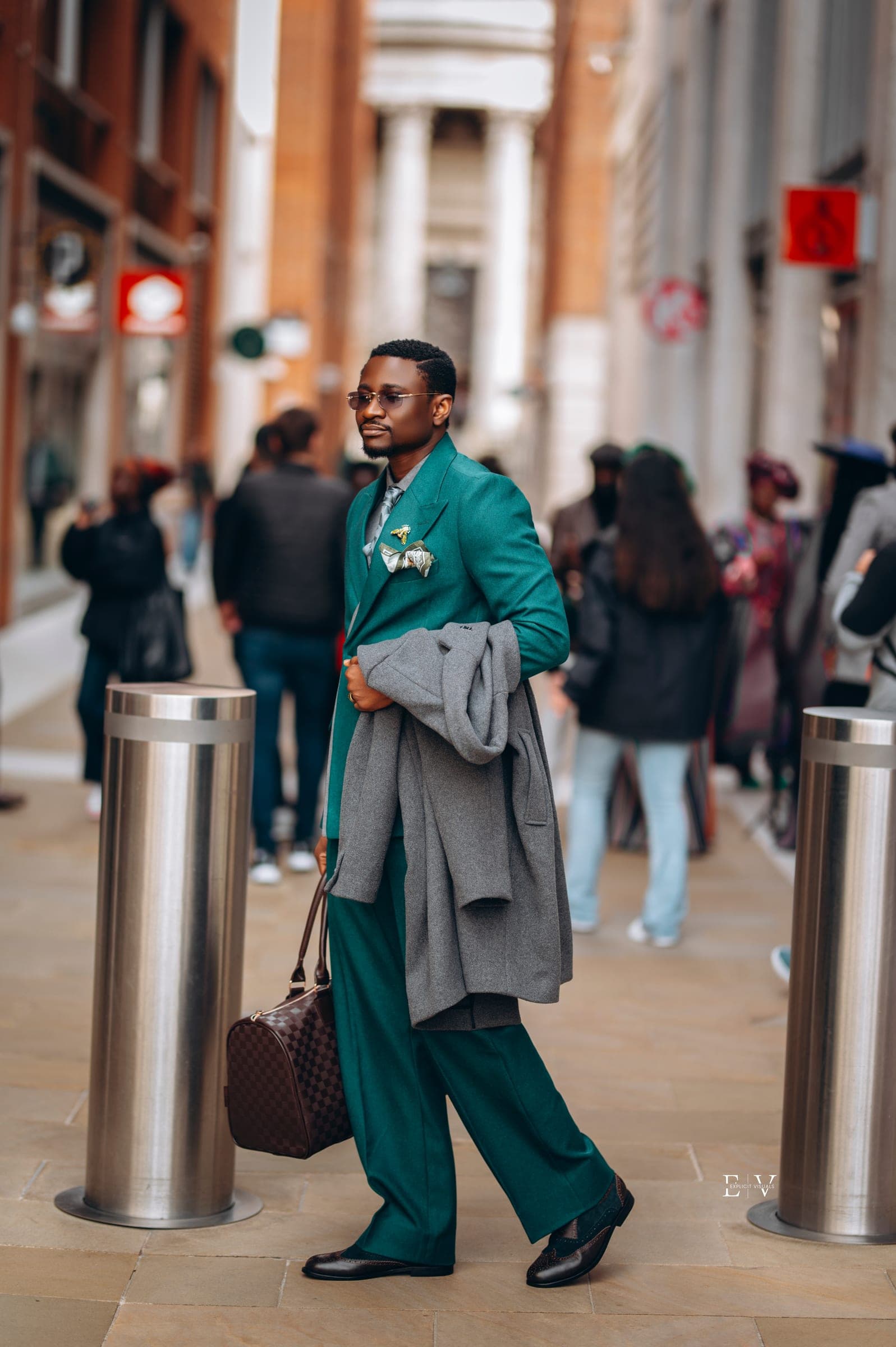 The same man walking the street in a full teal suit, a grey overcoat folded over one arm and a checked holdall in his hand.