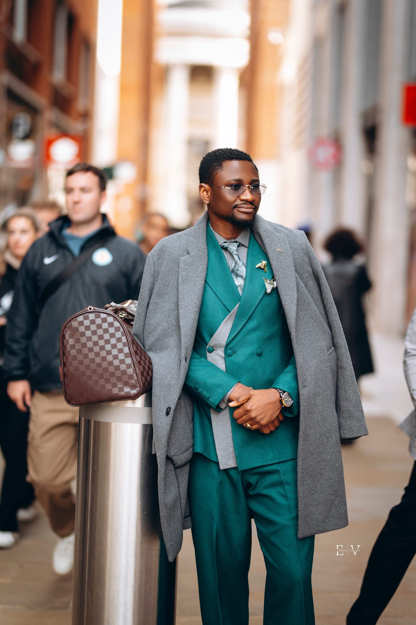 Standing still with the grey overcoat now across his shoulders, hands clasped, the teal three-piece beneath it.