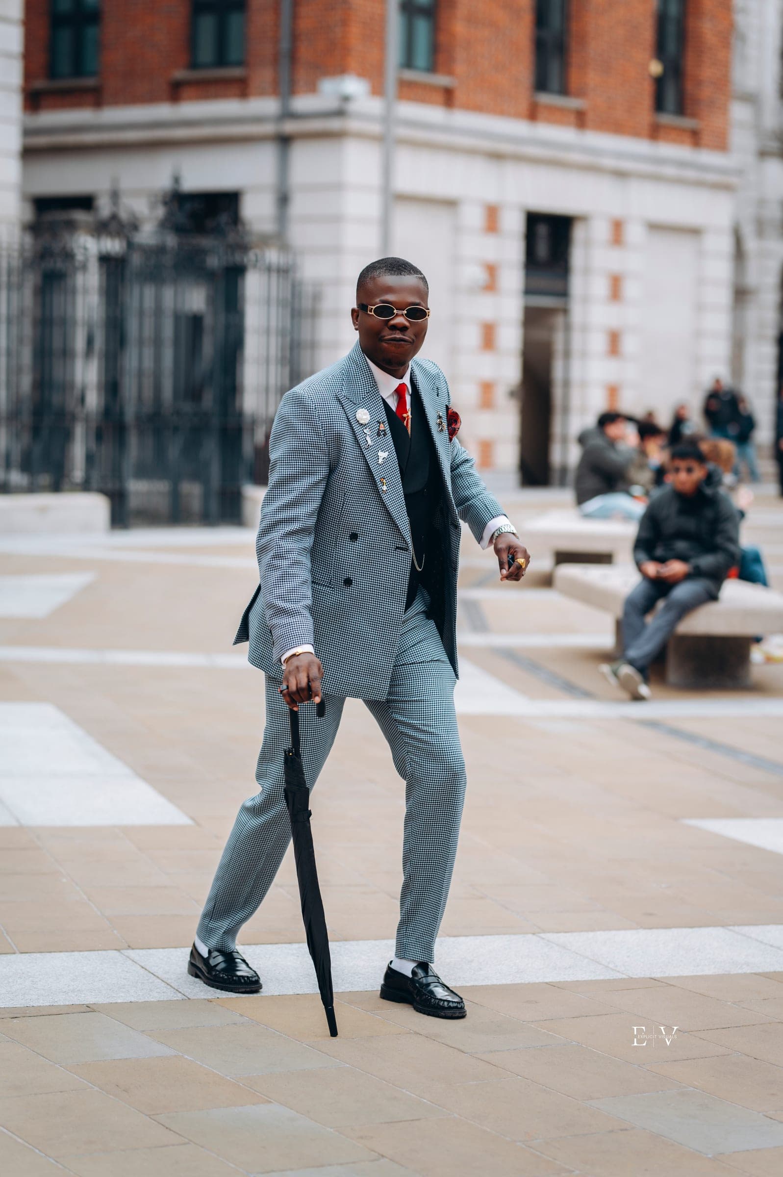 A man in a grey houndstooth double-breasted suit with a red tie and boutonnière, leaning on a black umbrella mid-step on the street.