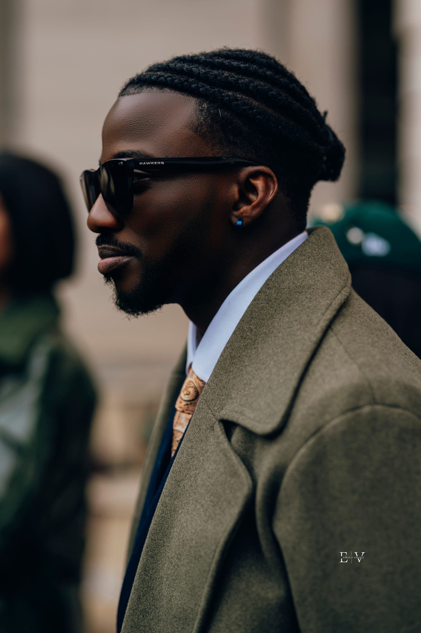 A profile portrait of a man in an olive overcoat and dark sunglasses, braids close to the head and a patterned tie against a white collar.