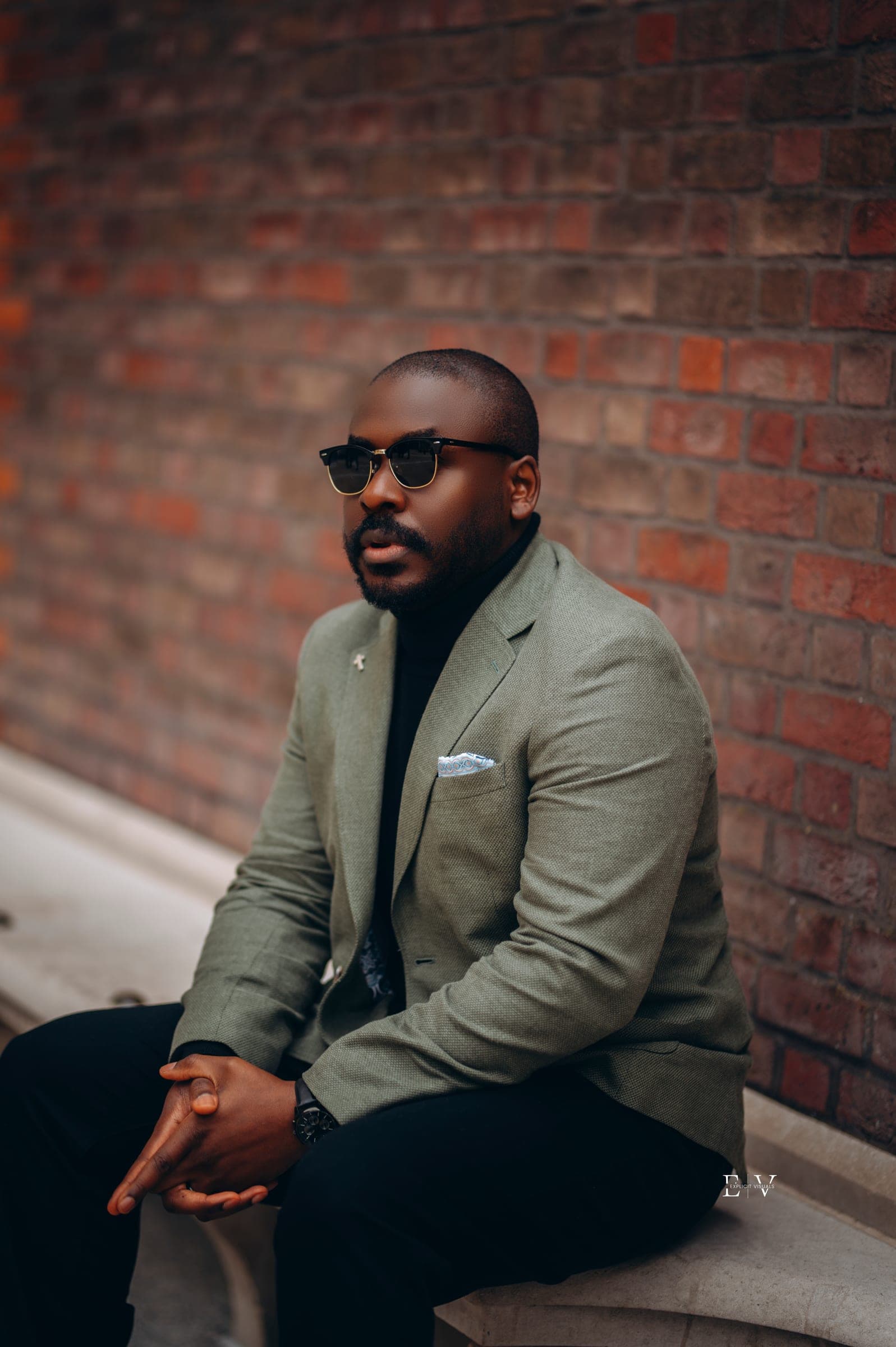 A man in a sage linen blazer and black roll neck sitting forward on a stone ledge against a red brick wall, hands loosely together.