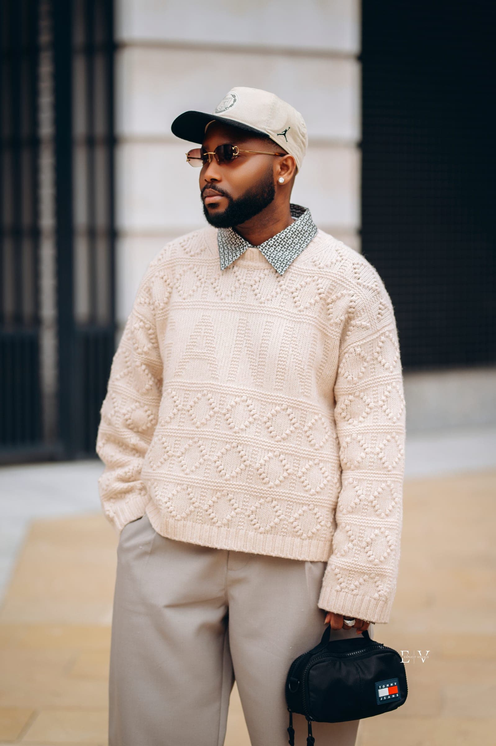 A man in a heavy cream cable knit worn over a sequinned collar, a cream cap and tinted glasses, a small black bag hanging from one hand.