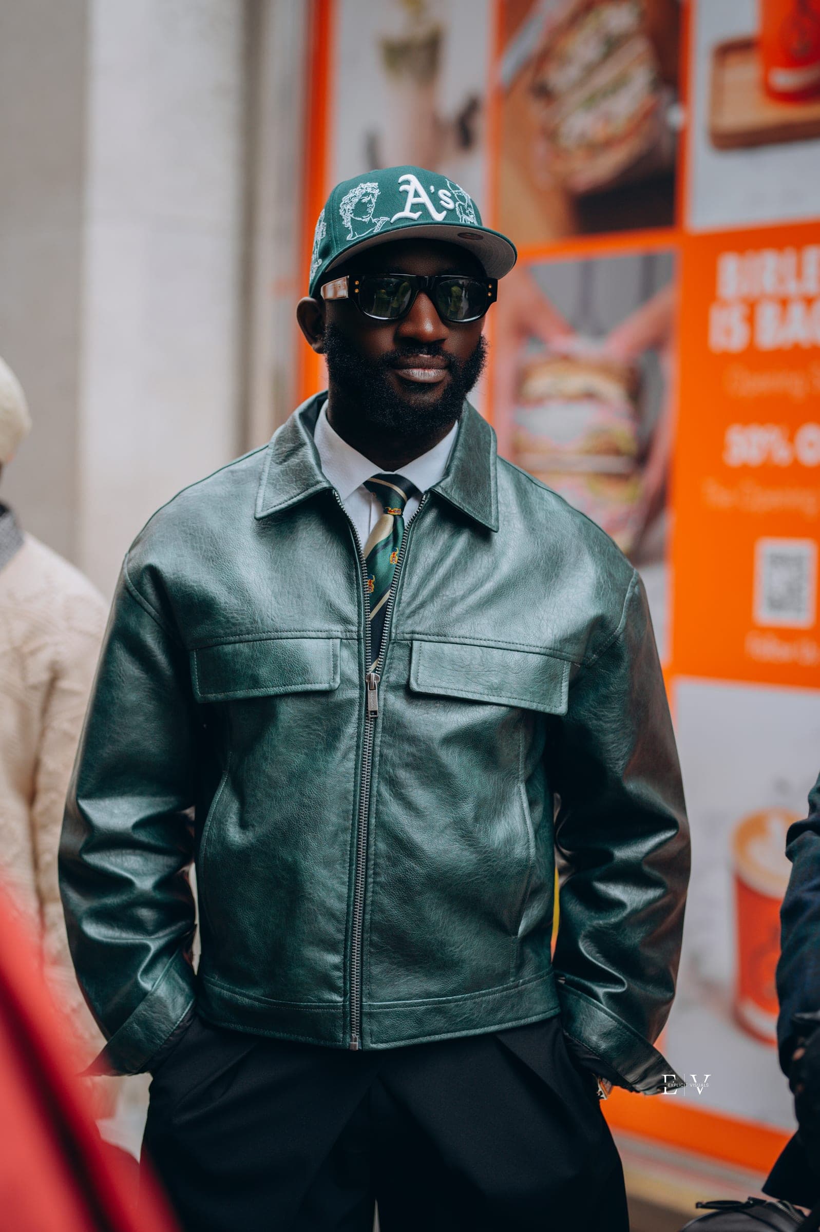 A man in a bottle green leather blouson over a shirt and tie, a green lettered ballcap and dark sunglasses, an orange shopfront poster behind him.