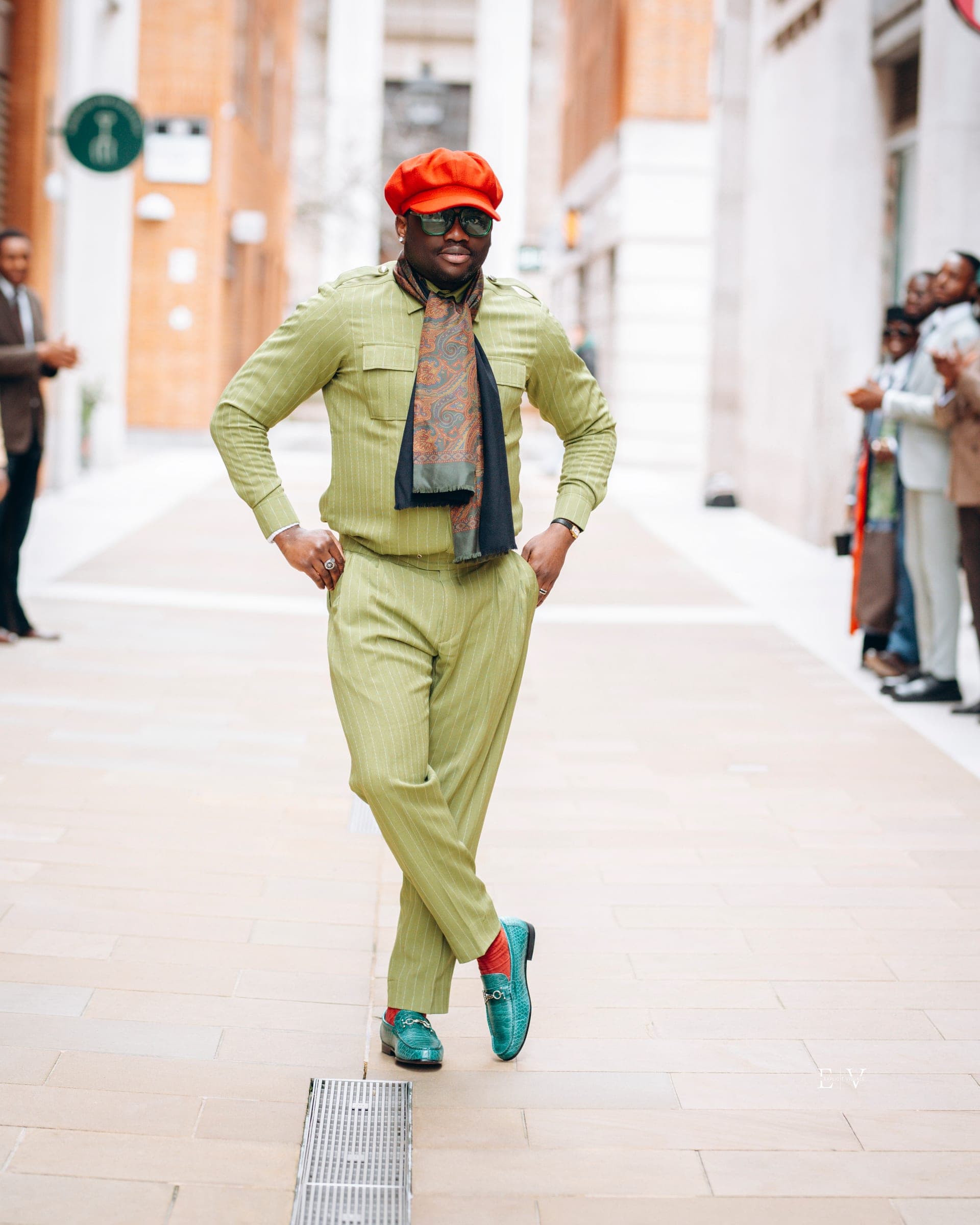 A man in a pistachio green shirt and trouser set with a printed scarf and an orange flat cap, mid-stride with teal and red loafers.