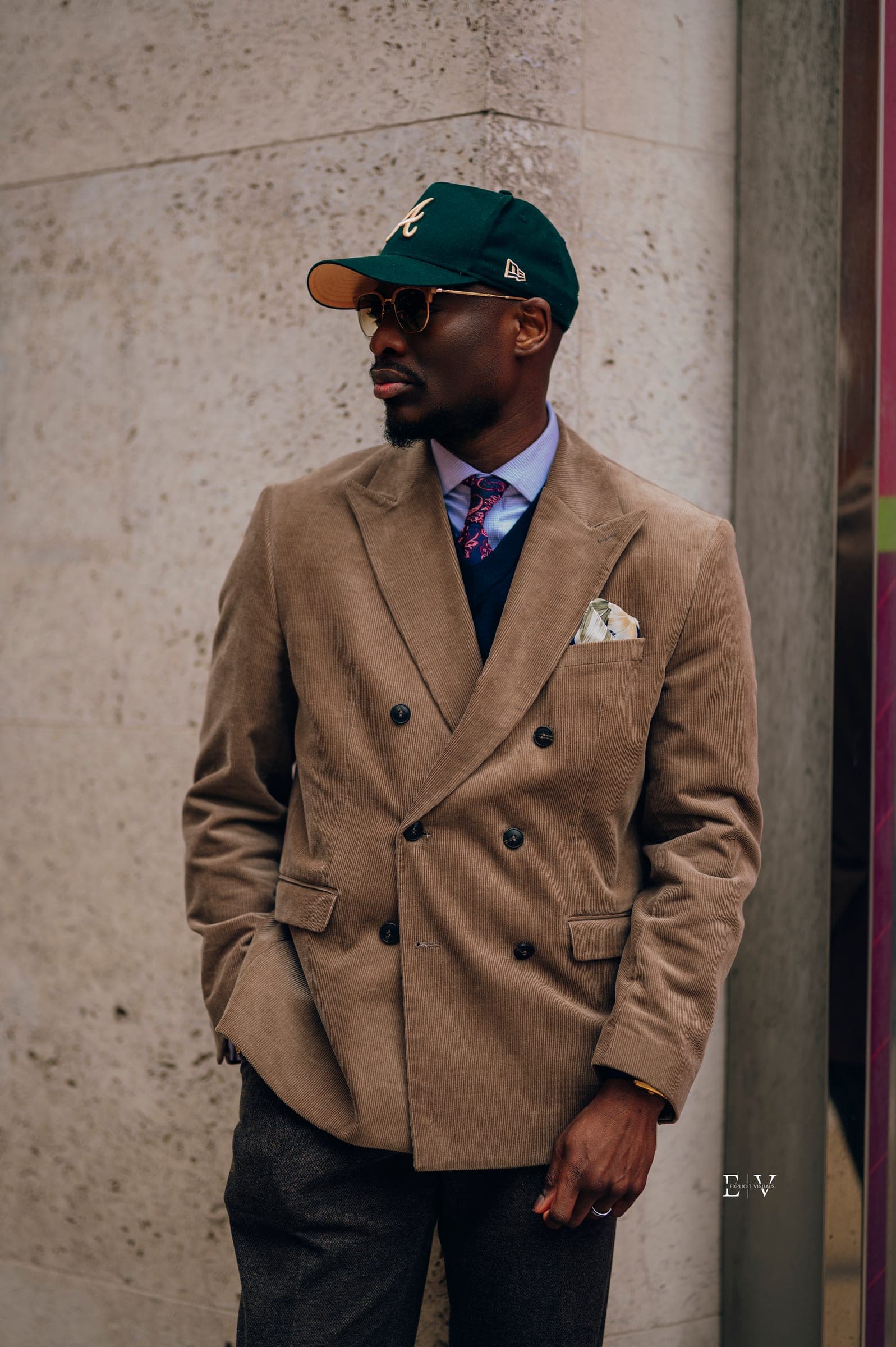 A man in a tan corduroy double-breasted blazer over navy, a floral tie and a white pocket square, wearing a green baseball cap.