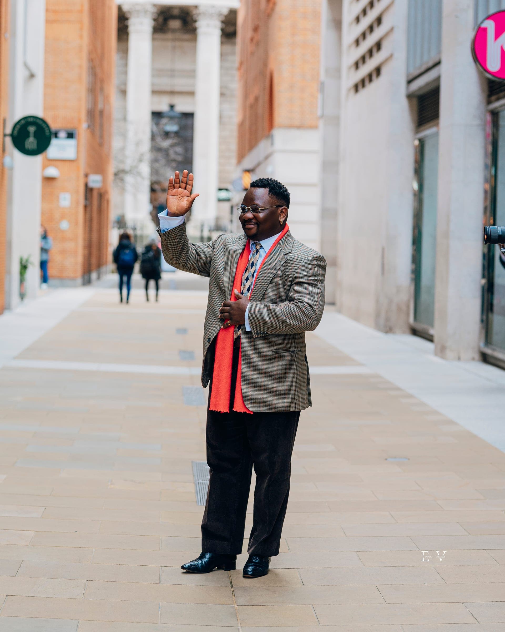 A man in a brown check blazer over a red shirt raising a hand to wave, alone in the middle of a pedestrian street.