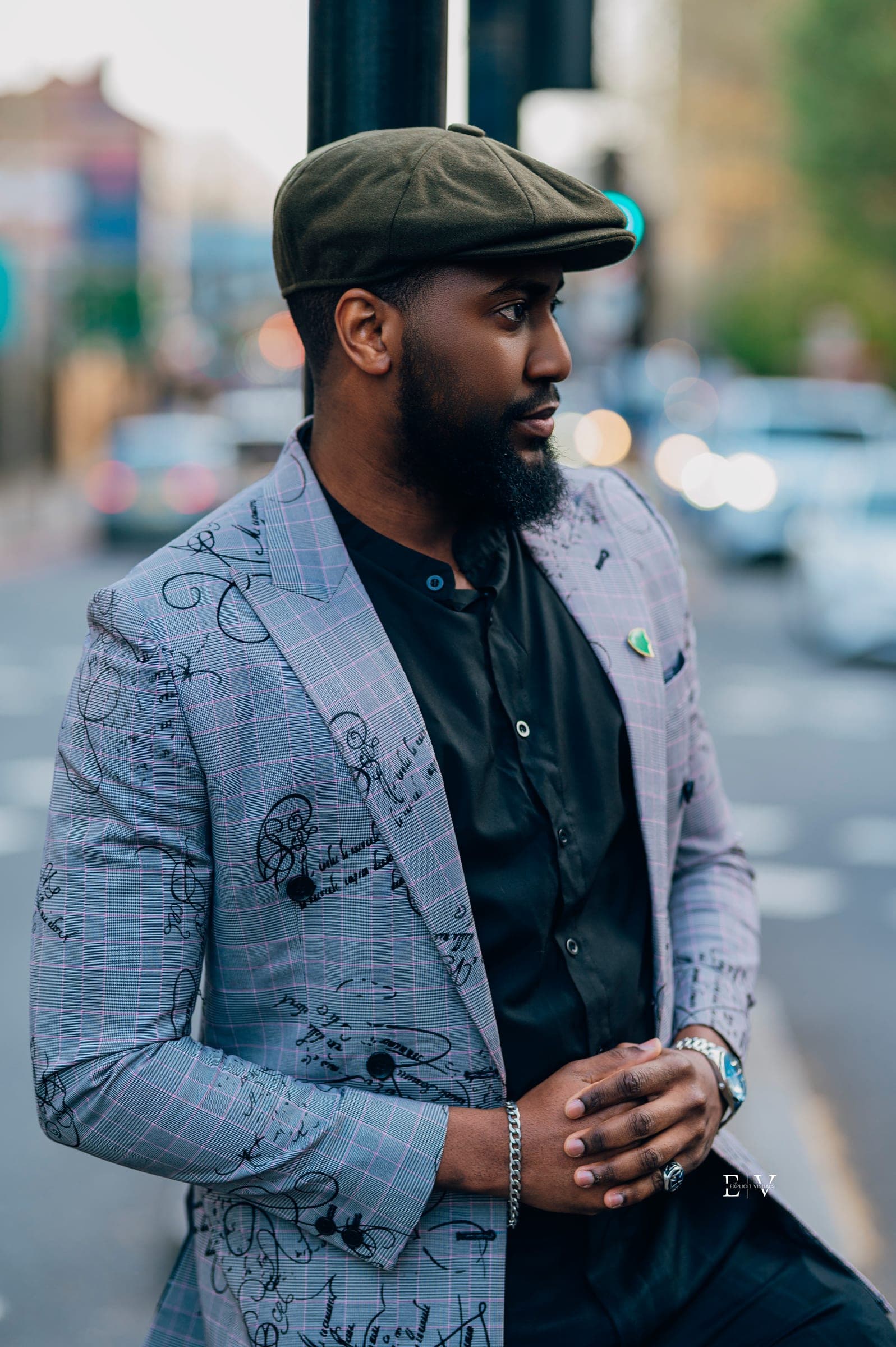 A man in an olive flat cap and a pale checked blazer printed with line drawings, over a black shirt, standing at the kerb with traffic blurred behind him.