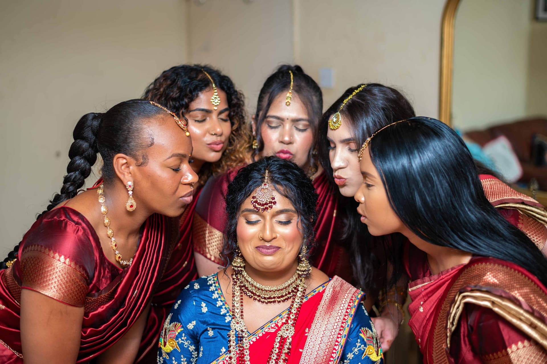 Five bridesmaids in deep red sarees lean in around the seated bride from every side, eyes closed, all mid-kiss.