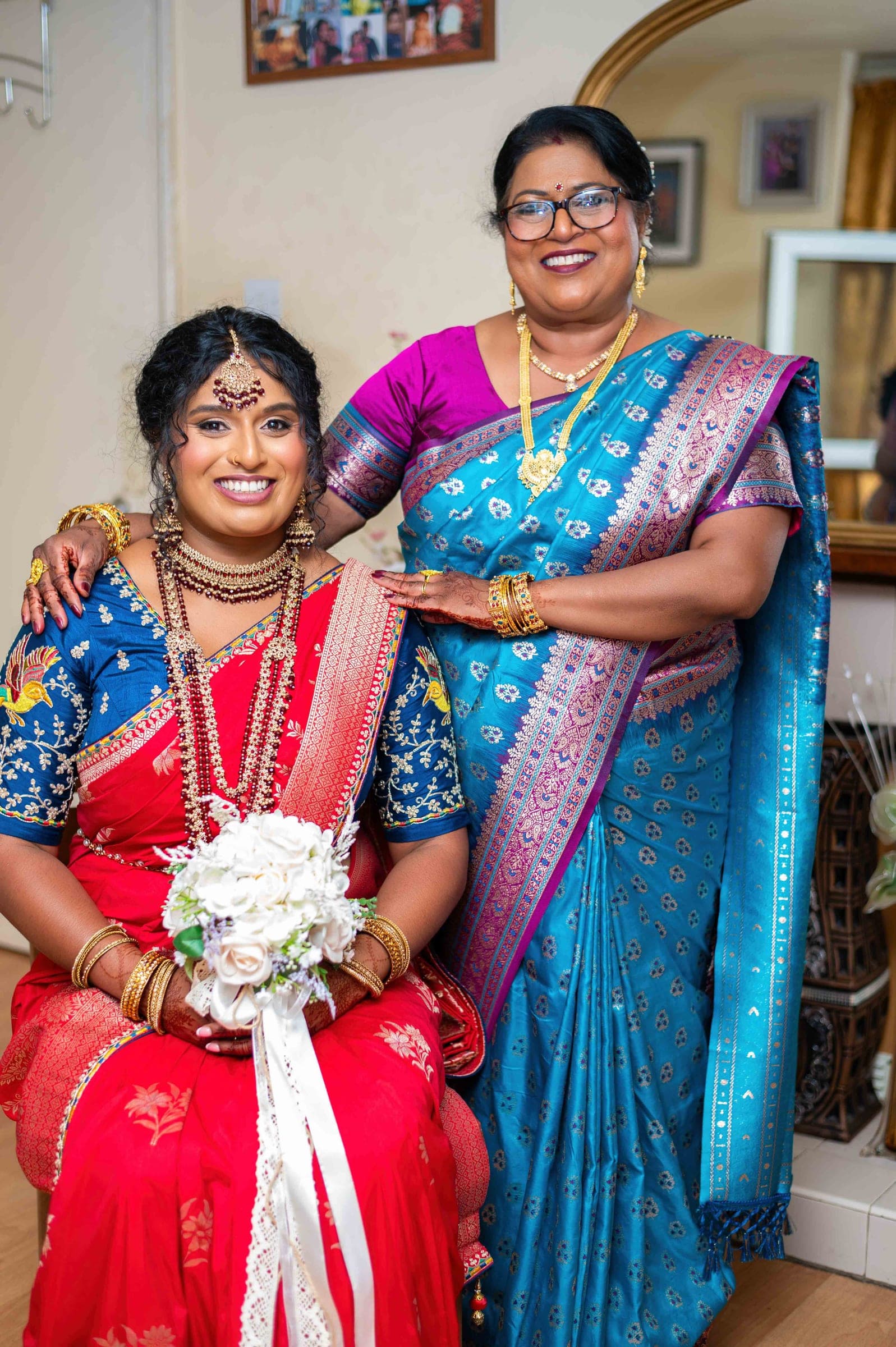 The bride seated beside her mother, who stands in a teal and magenta silk saree with a hand on her daughter's shoulder.
