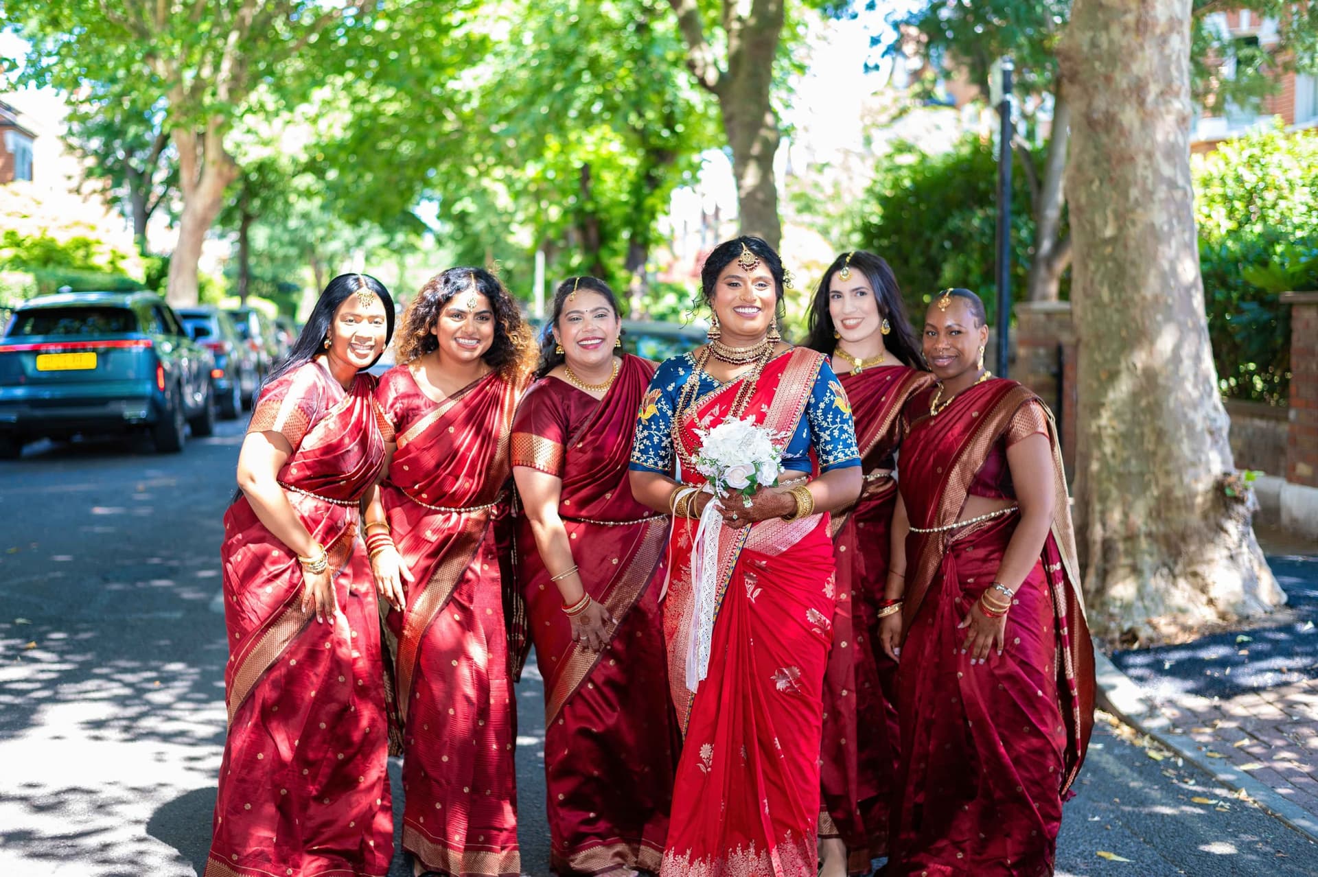 All six walking a residential pavement together, the bride centre in bright red, the bridesmaids in deeper red on either side.