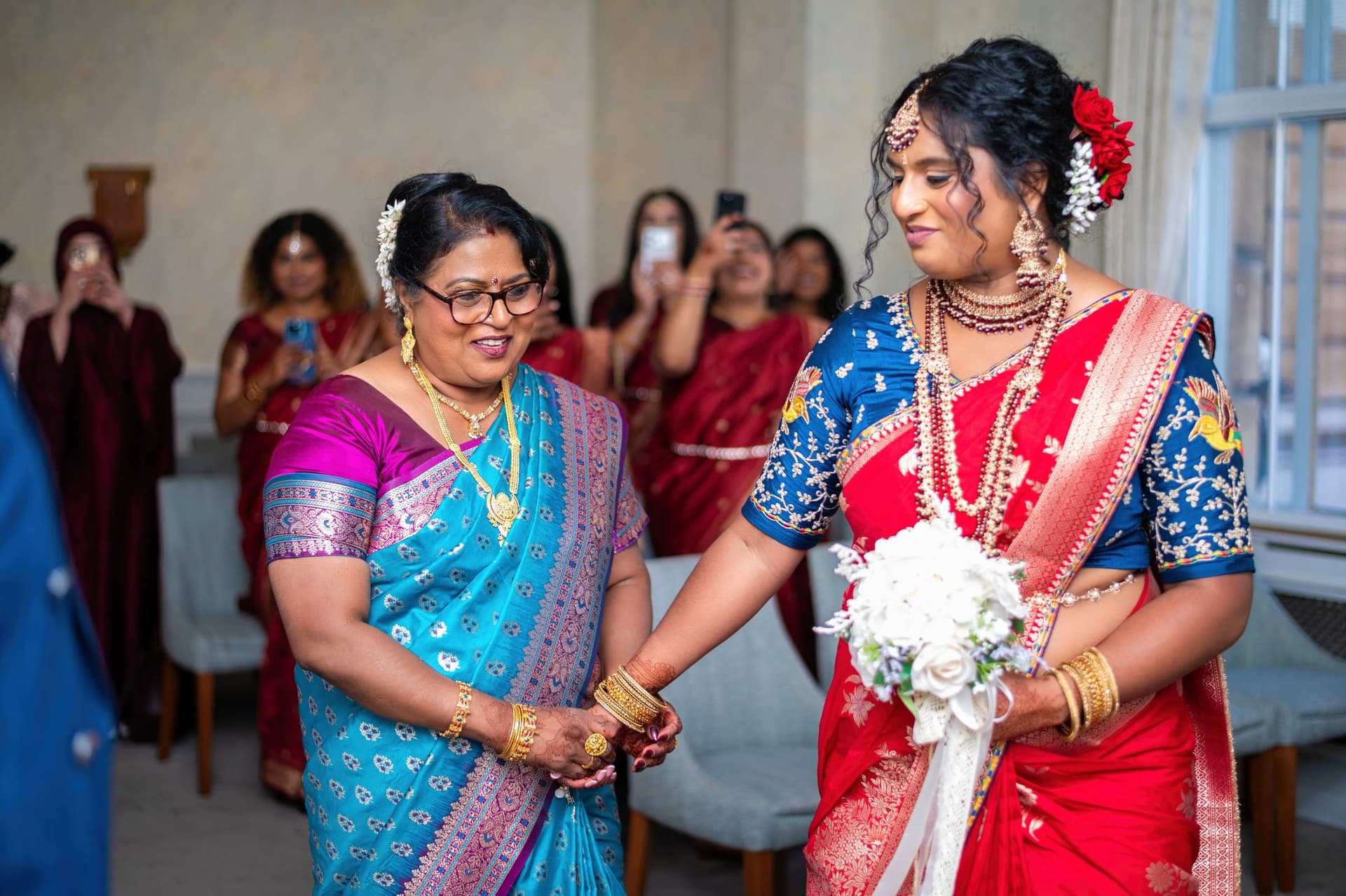 The bride walks into the ceremony room hand in hand with her mother, guests lifting phones on both sides.