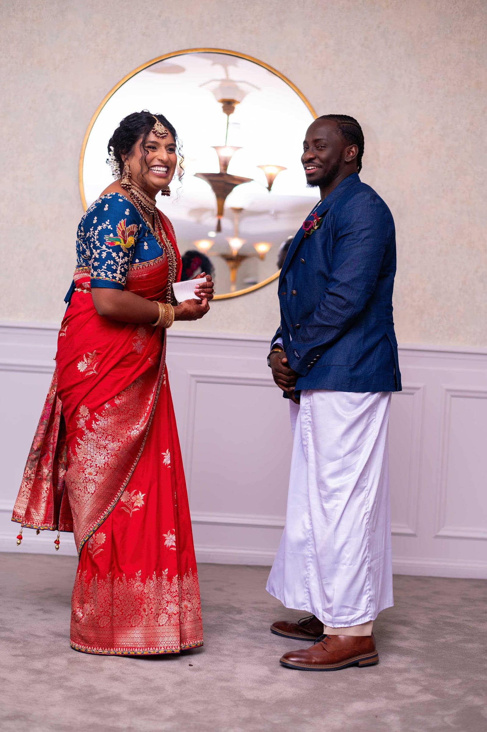 The two of them facing each other and laughing before a round gilt mirror, he in a blue denim blazer and white veshti.