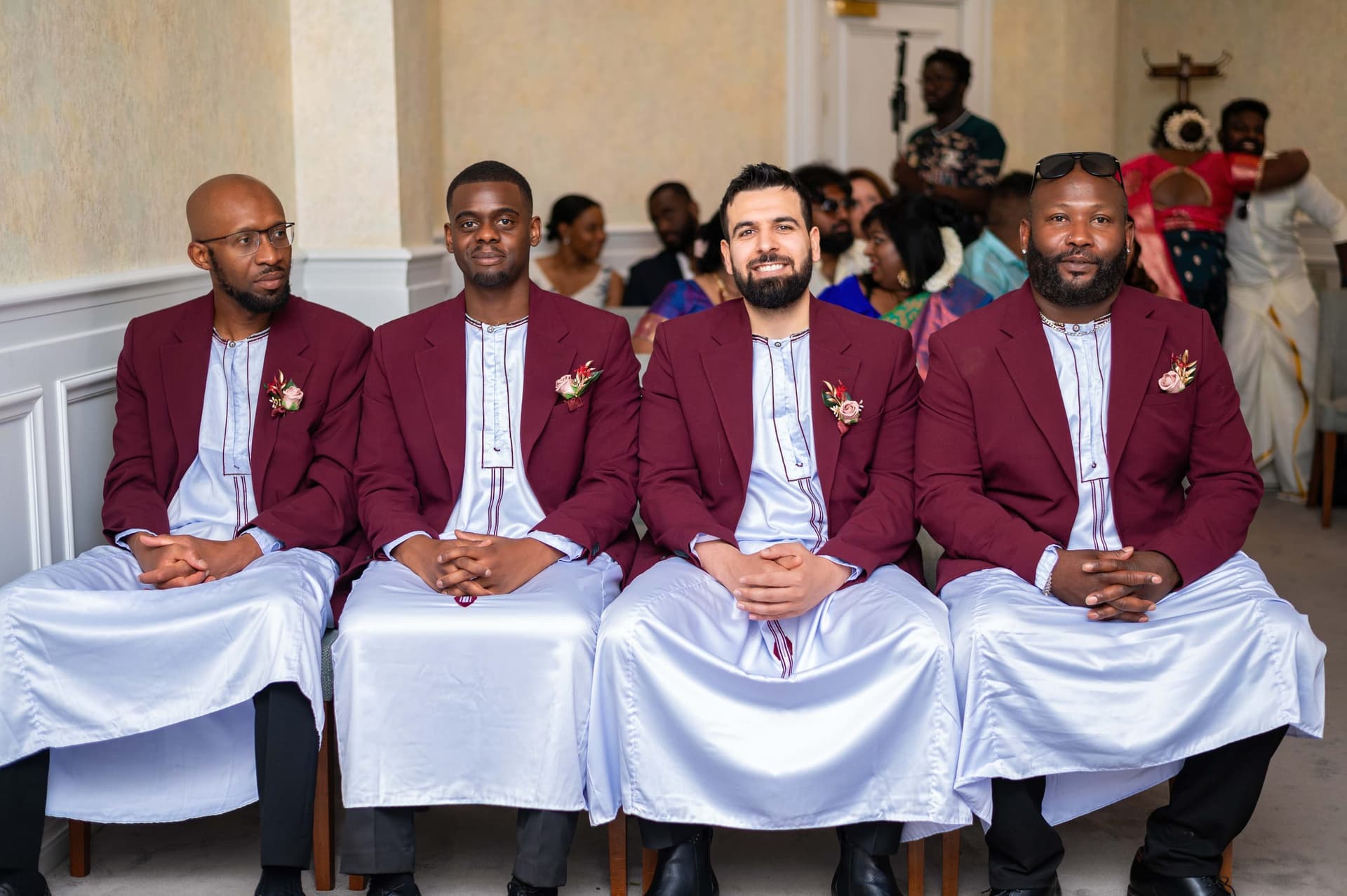 Four groomsmen seated in a row in burgundy blazers over pale blue kaftans, rose boutonnières pinned at the chest.