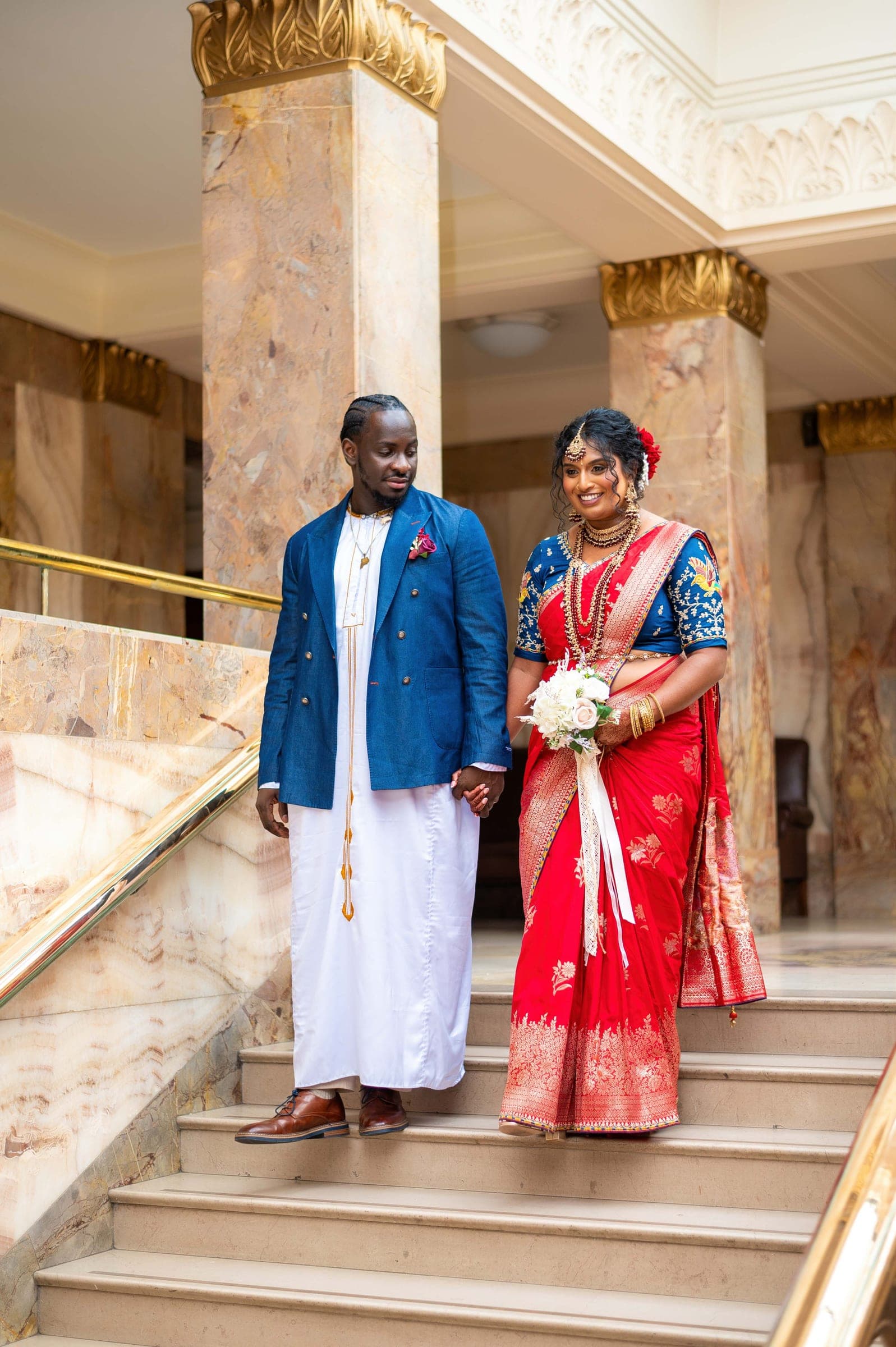 The couple stand hand in hand on a marble staircase between gilded columns.