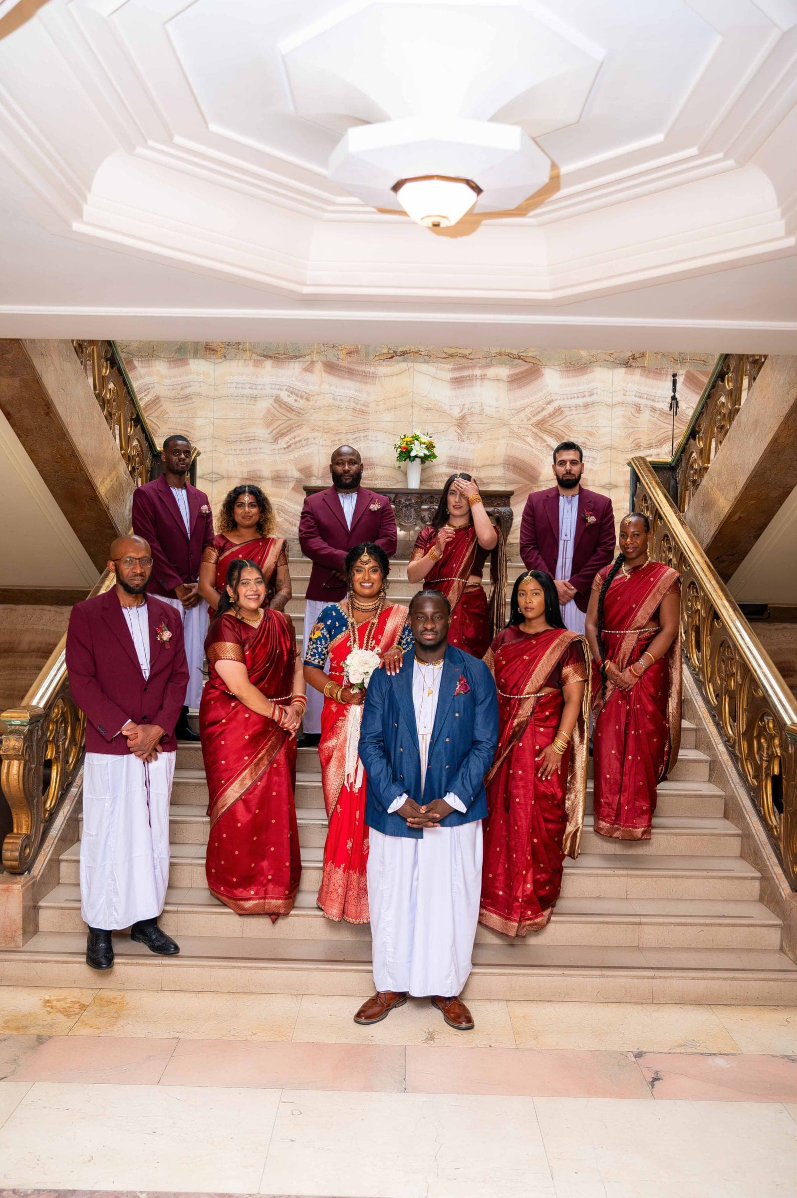 The whole bridal party arranged up the marble staircase — groomsmen in burgundy, bridesmaids in red, the couple at the centre.