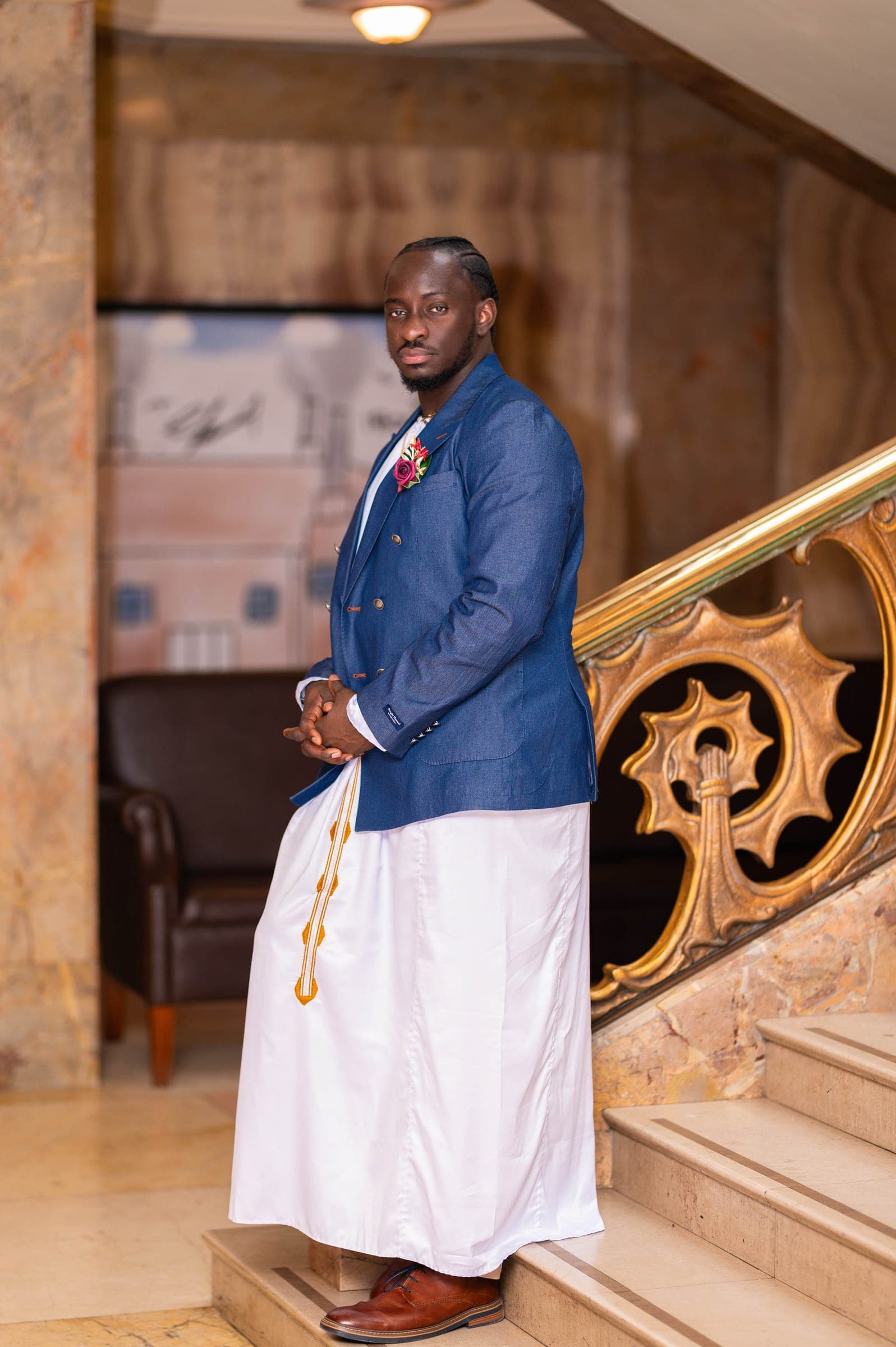 The groom full-length on the marble stairs beside the gilded banister, hands clasped.