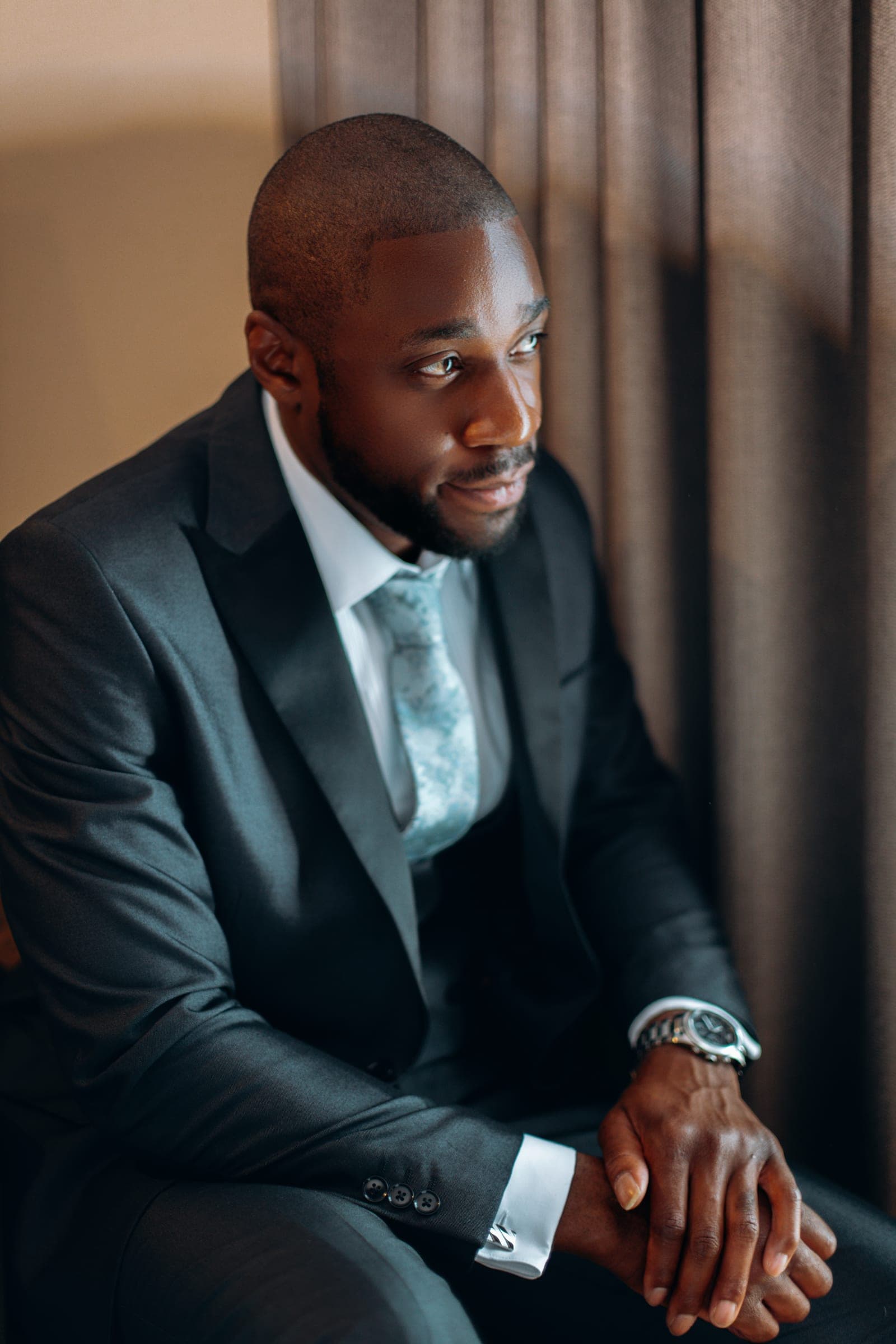 The groom seated in a charcoal three-piece suit and a pale patterned tie, looking away from the camera.