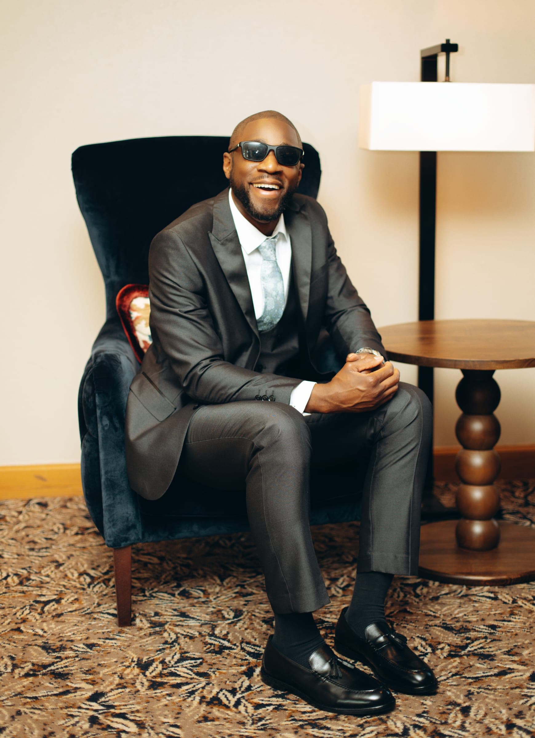 The groom laughing in a navy velvet armchair, sunglasses on, one ankle across his knee.