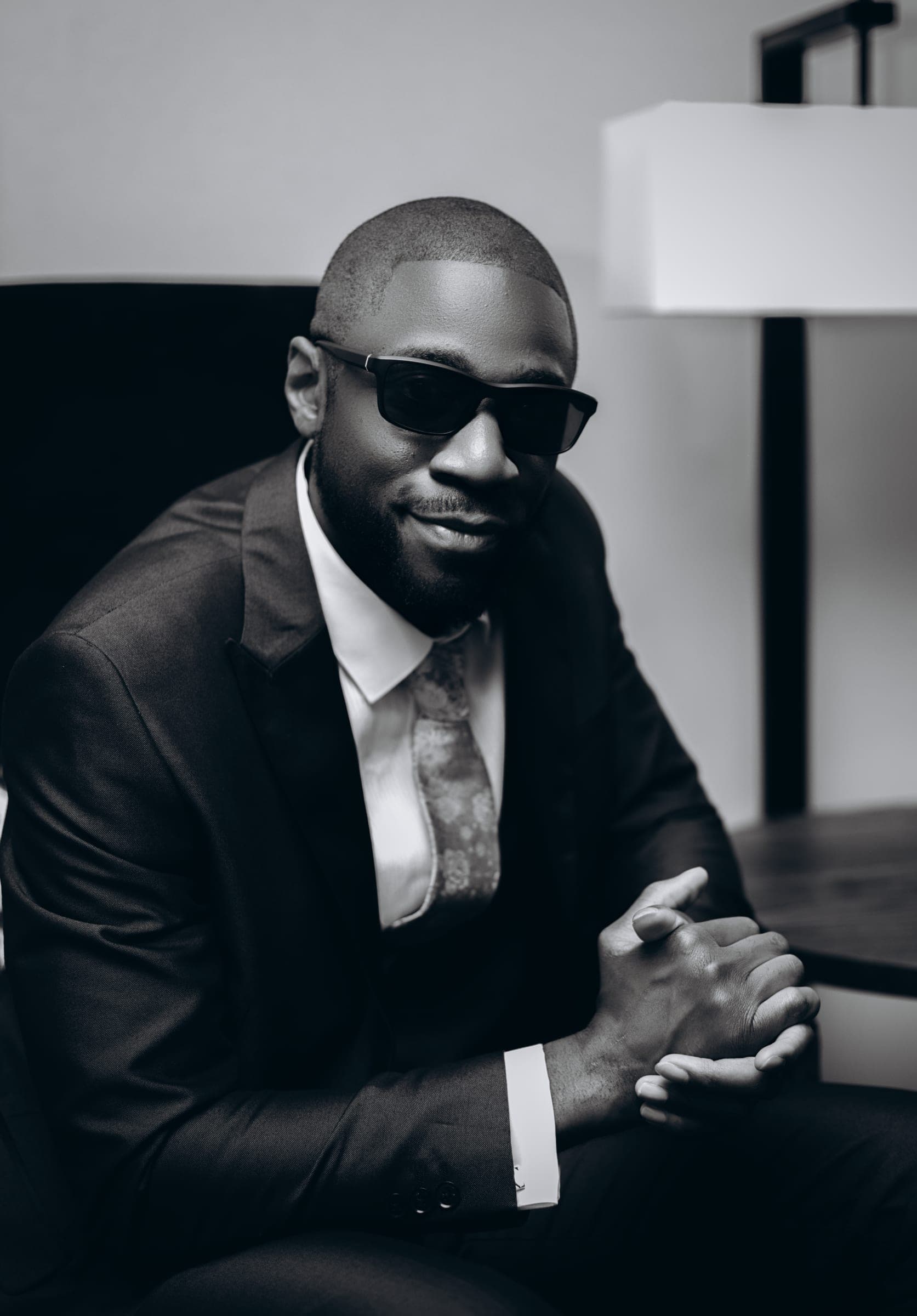 A tight black and white portrait, dark glasses on and hands clasped in front of him.