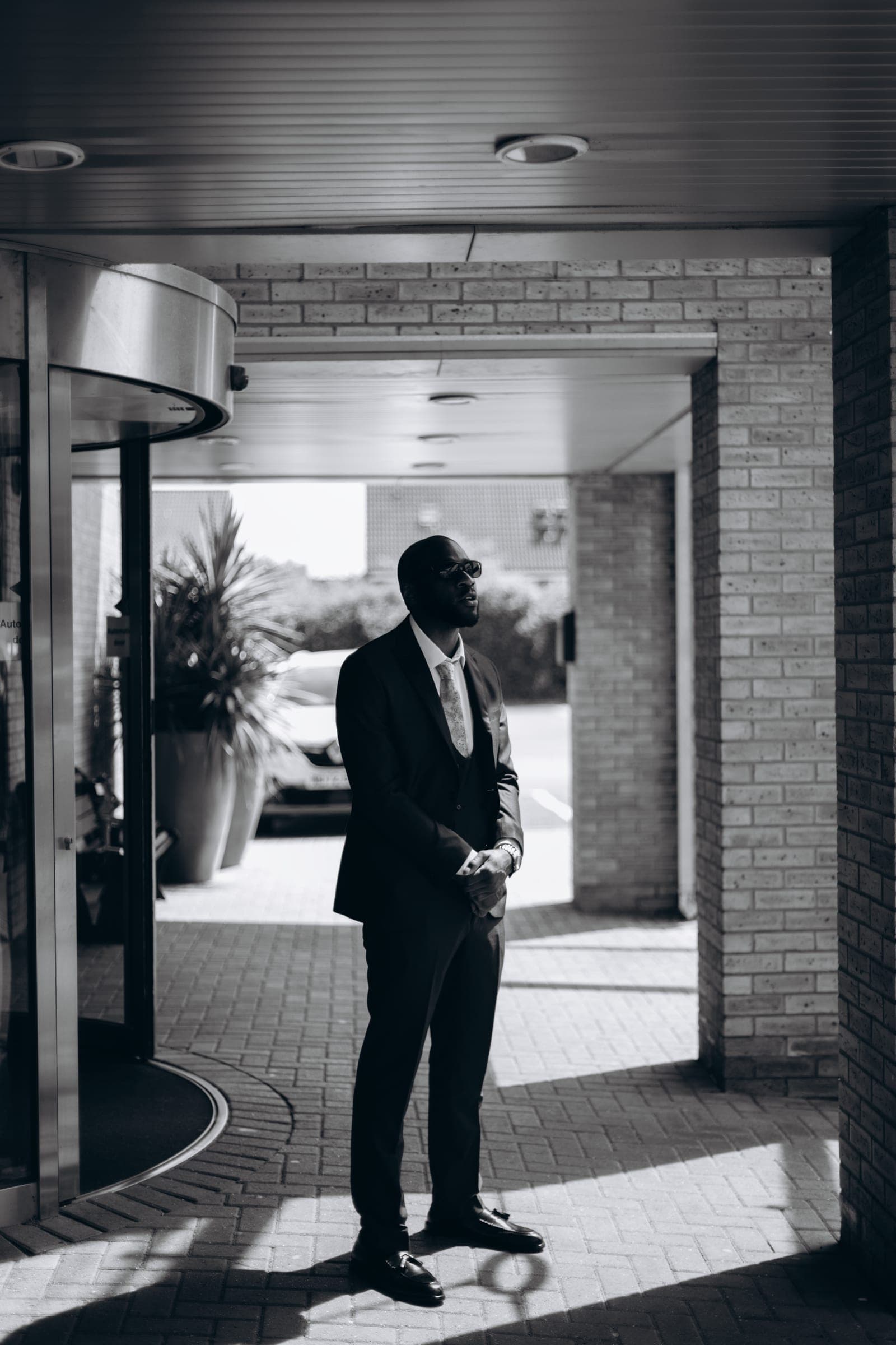In black and white, the groom stands alone outside the hotel entrance, light falling across the paving.