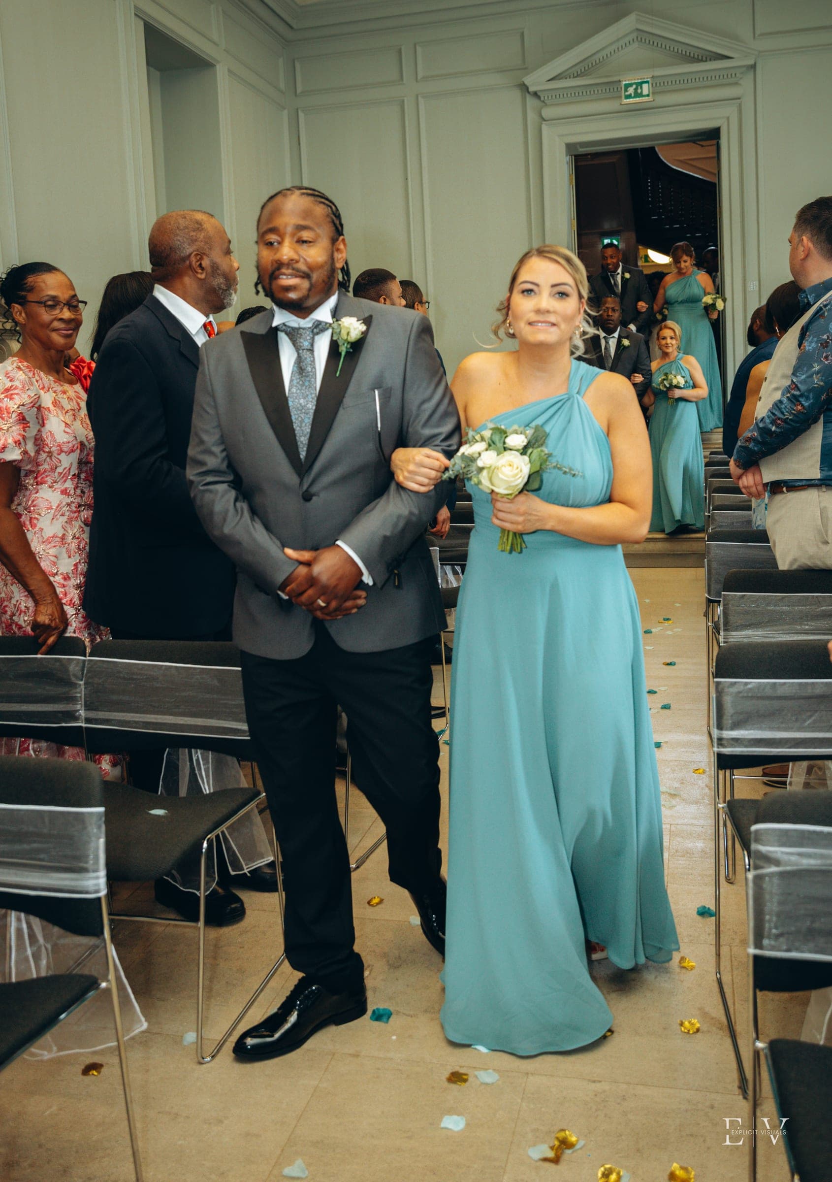 The same pair further down the aisle, petals and confetti already scattered across the floor.