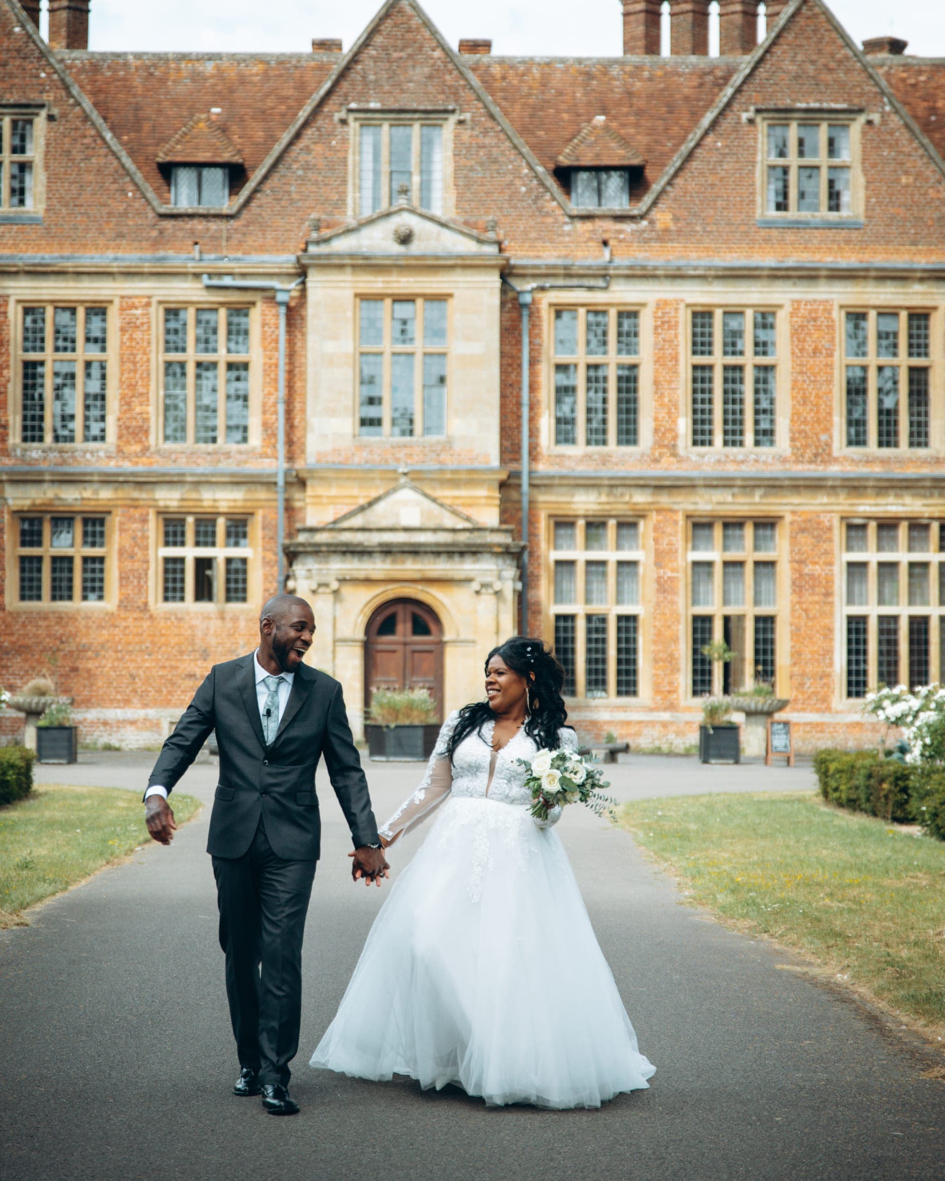 The couple walk hand in hand up the drive with a red brick Jacobean manor house filling the frame behind them.