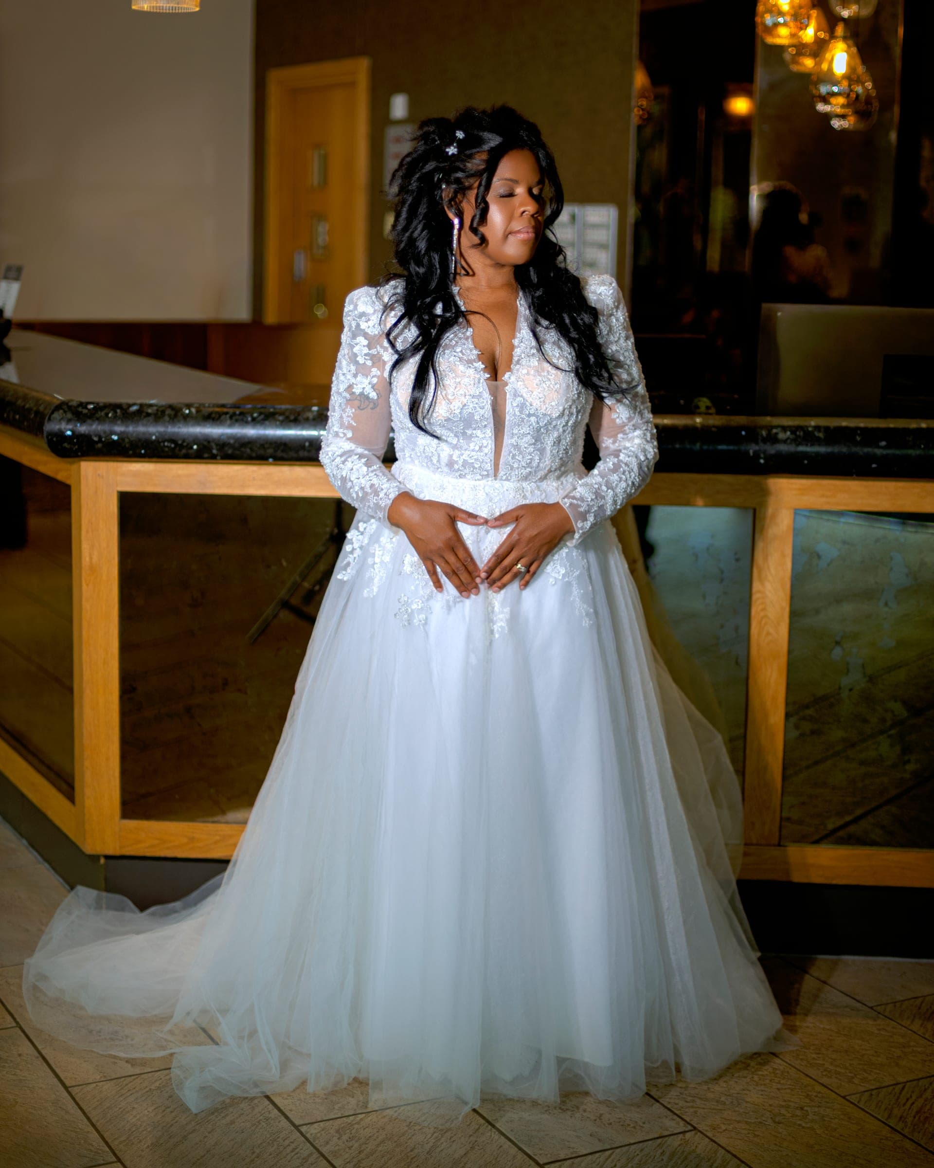 The bride indoors against a glass balustrade, head tipped, hands together.