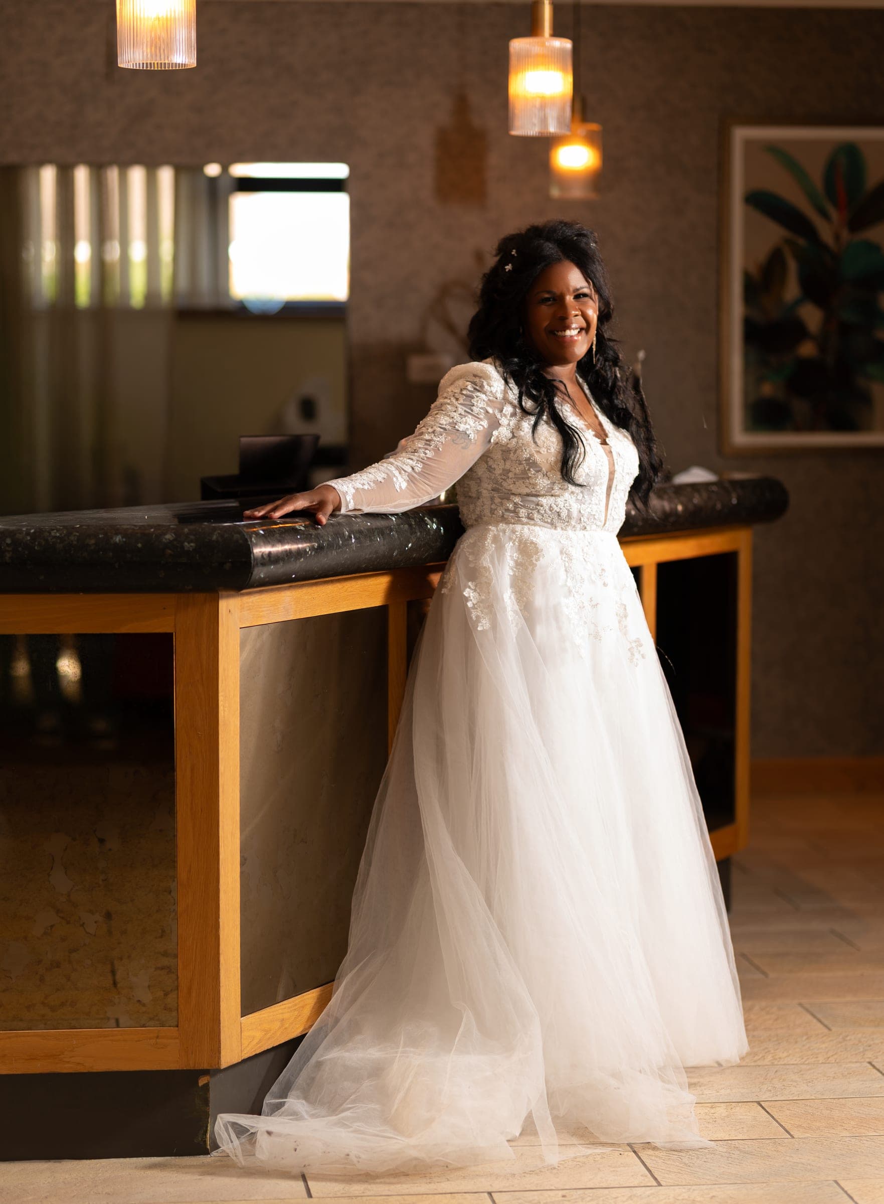 The bride leaning against a dark stone bar top, laughing, pendant lights warm overhead.